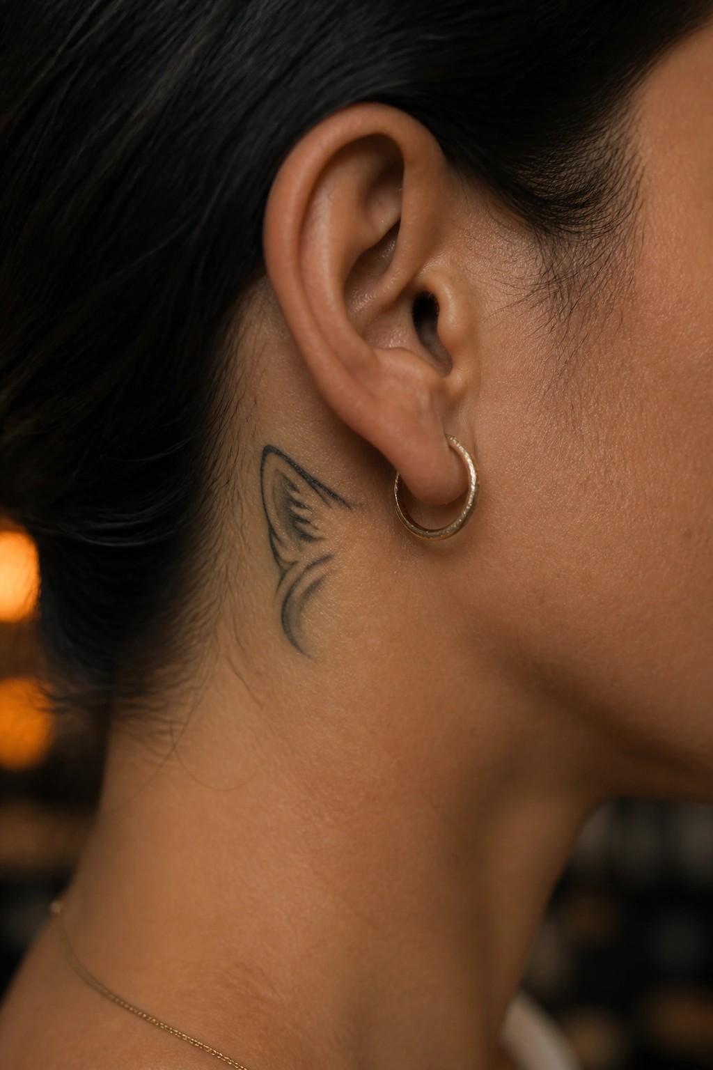 Close up of a small black line art wolf ear tattoo placed behind a woman's ear on her neck next to a thin gold hoop earring.