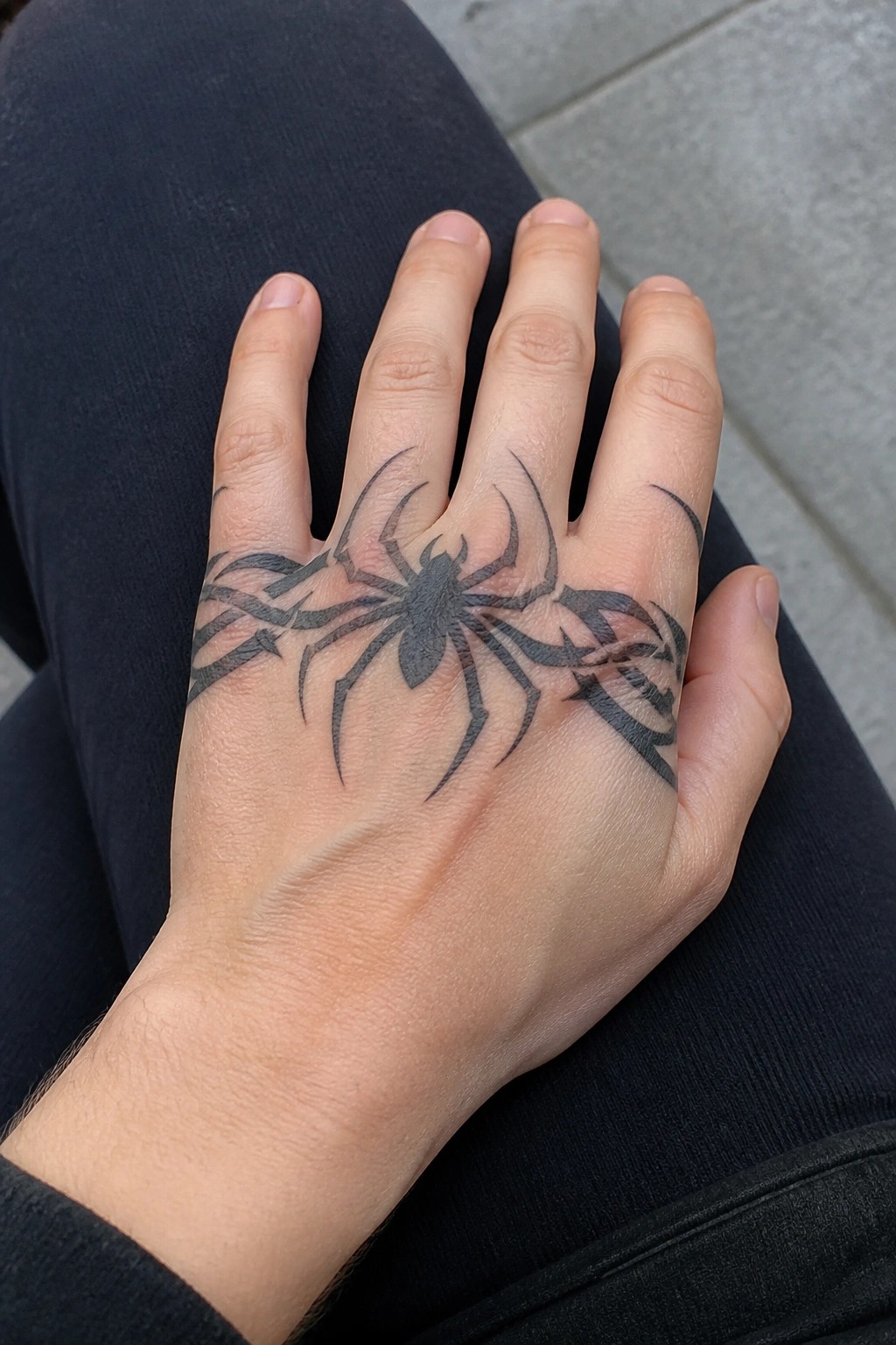 Hand with large black spider tattoo resting on dark pants over gray pavement