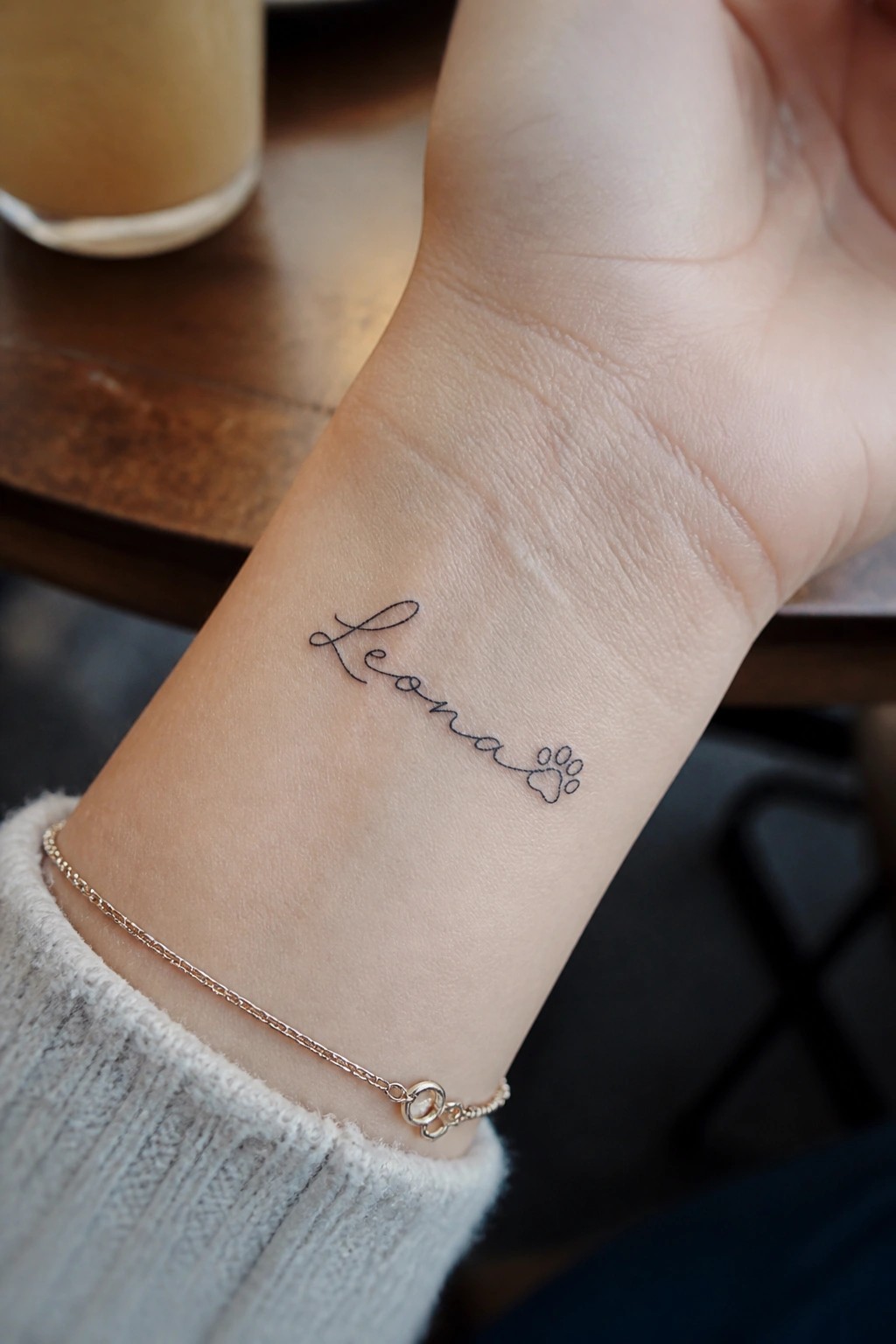 Fine line cursive tattoo spelling Leona with a small paw print on the inner wrist