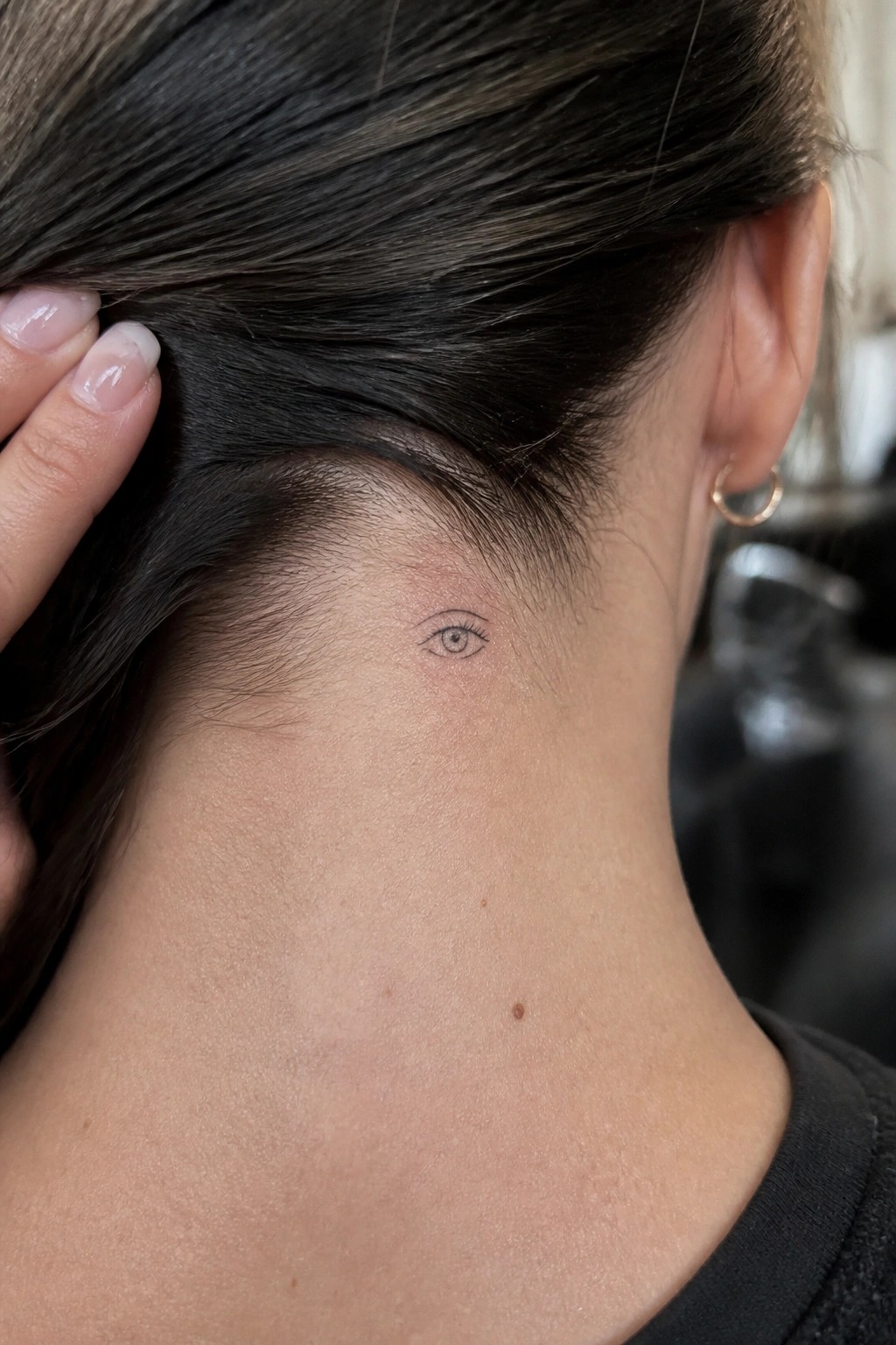 A small fine line eye tattoo positioned just below the hairline on the back of a woman's neck.