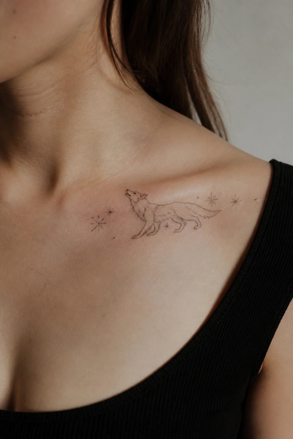 Fine line she wolf tattoo on the collarbone with small stars around it, shown on light skin in a close up photo.