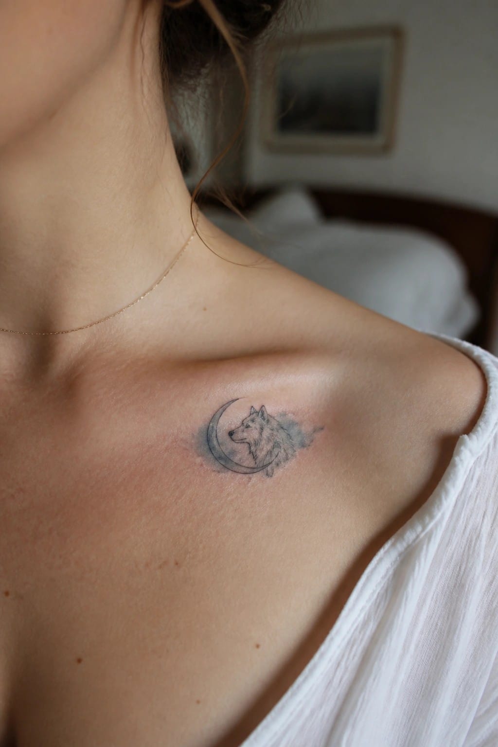 Small fine line tattoo of a wolf head inside a crescent moon on the collarbone with soft grey shading.