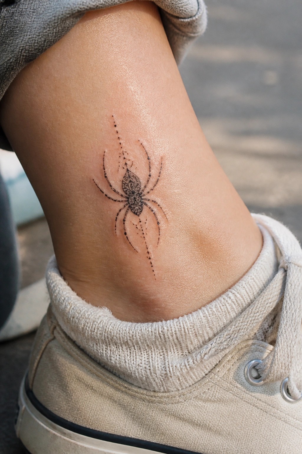 Small fine line spider tattoo on the ankle with dotted accents and an ornate body pattern above a white sneaker.