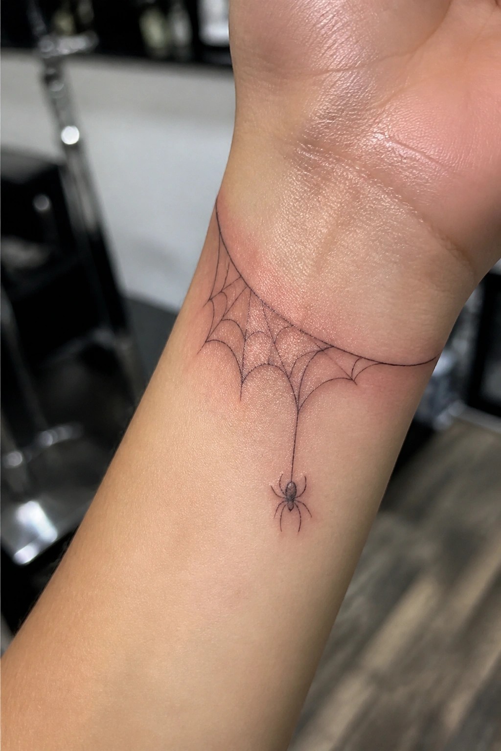 Delicate black spider web tattoo with hanging spider on wrist.