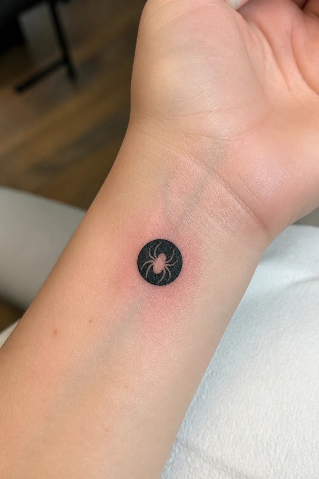 Small black circular spider tattoo on reddened inner wrist skin