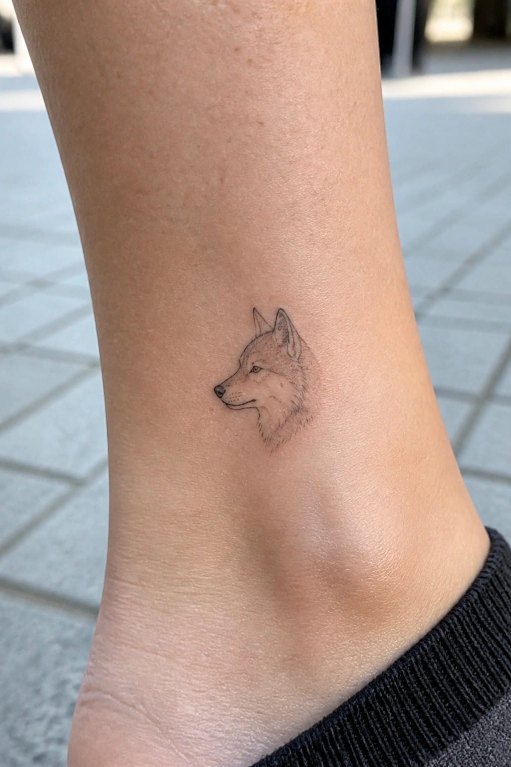 Black ink wolf head tattoo on light skin ankle above black cuff