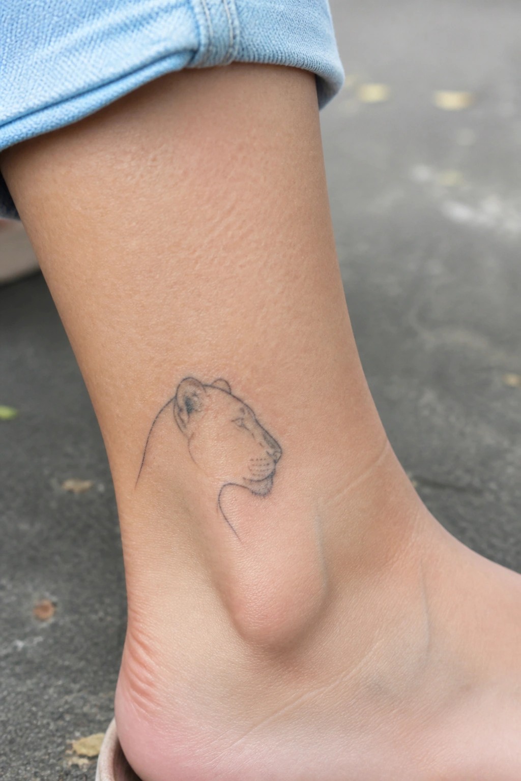 Cuffed light blue jeans frame a minimalist lioness tattoo on bare ankle.