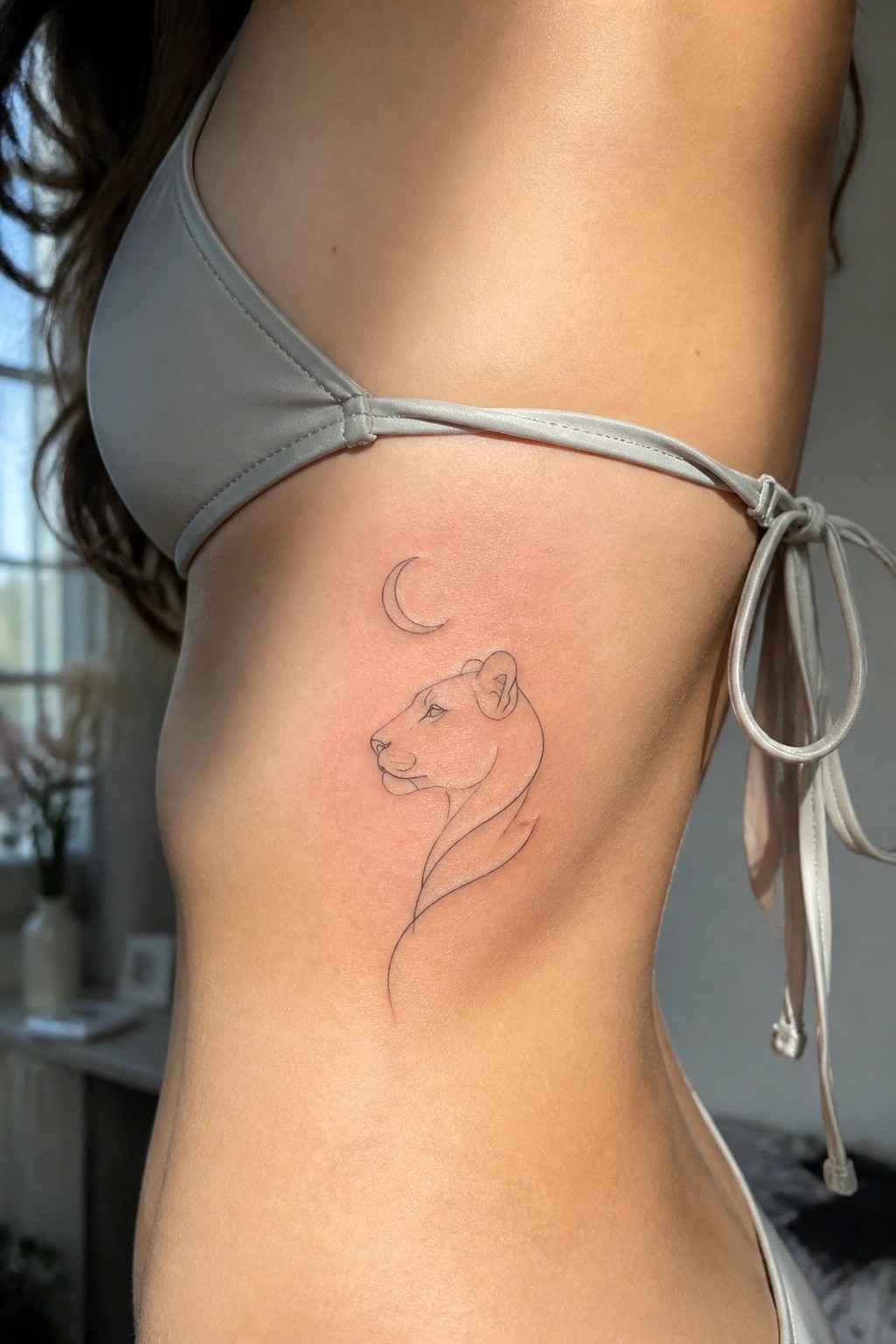 Woman's torso with fresh minimalist lioness and crescent moon line tattoo under light gray bikini.
