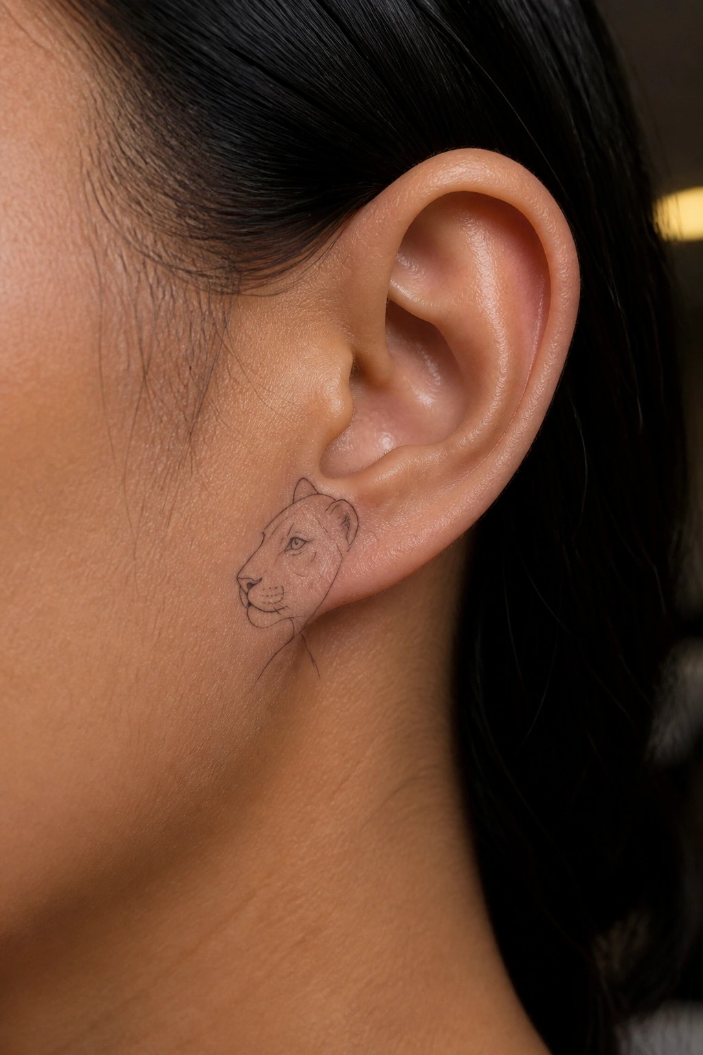 Small line-art lioness tattoo behind a person's ear amid dark hair.