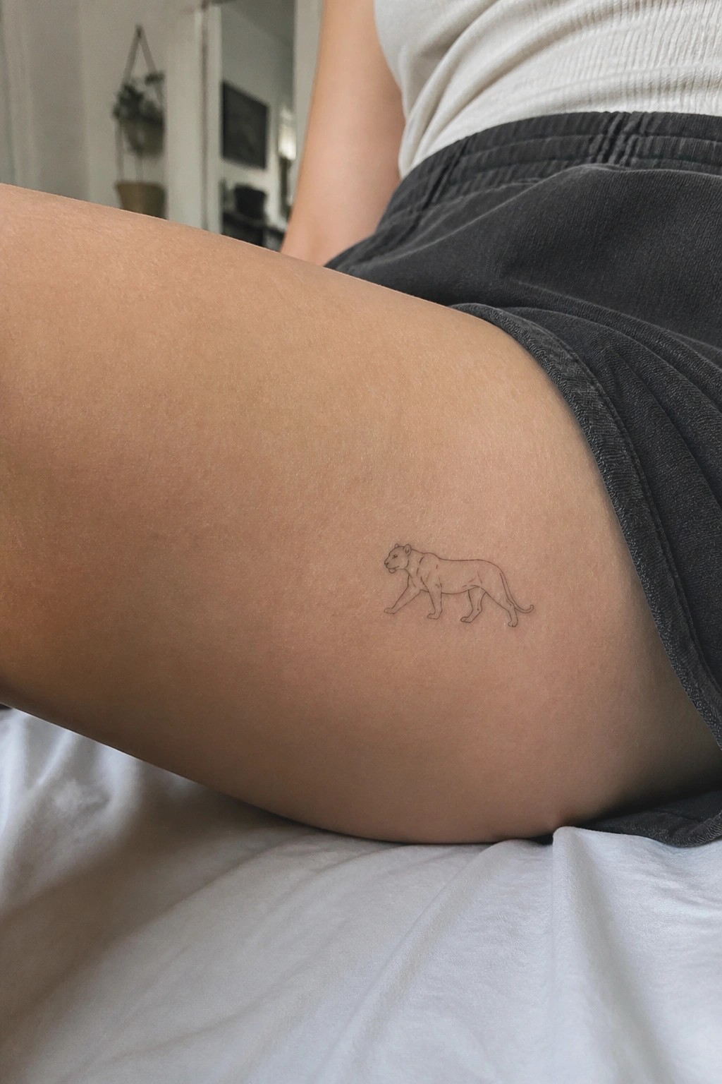 Small fine-line walking lioness tattoo on thigh beside grey shorts and white top