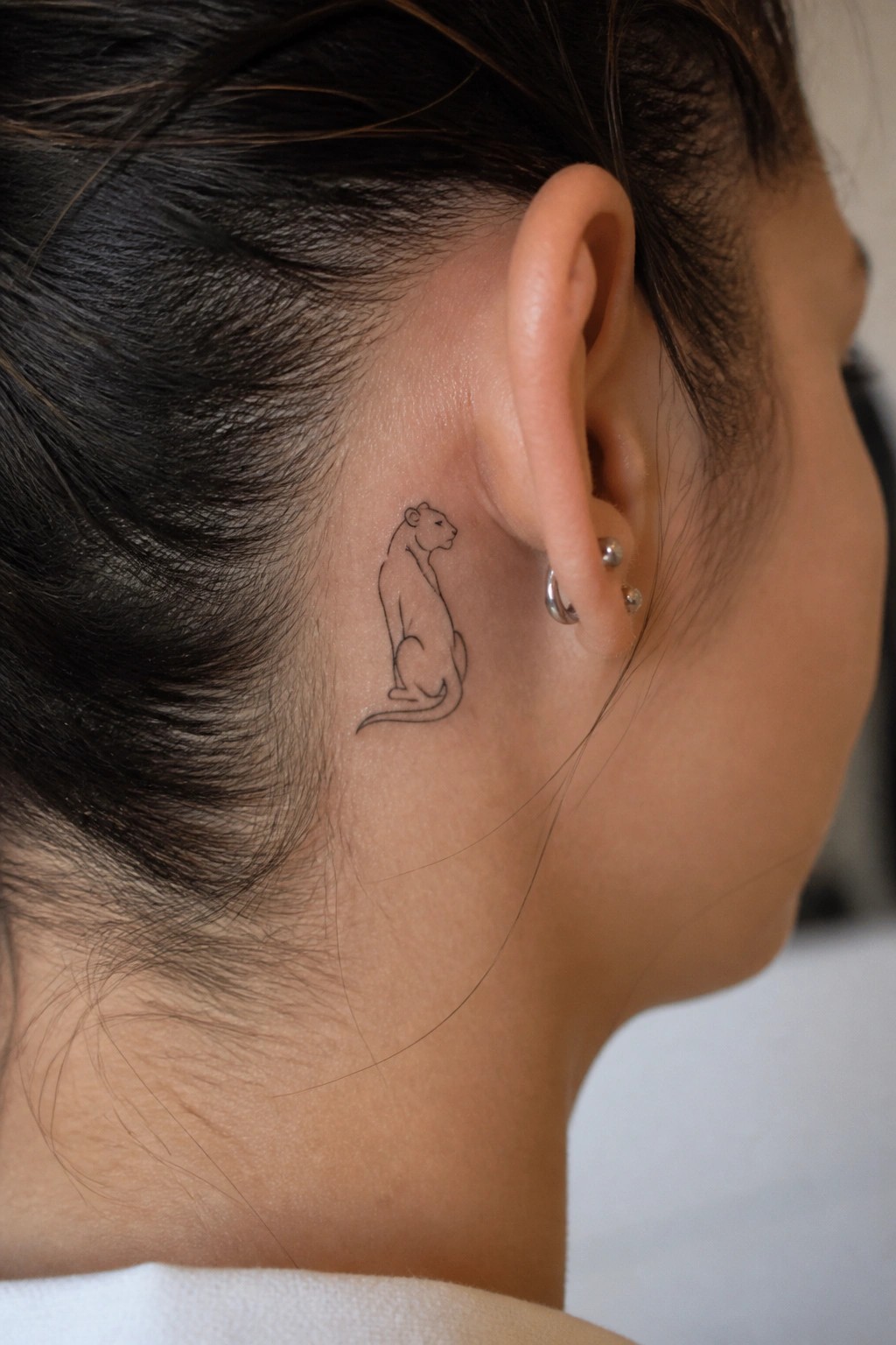 Small minimalist line art lioness tattoo placed behind a woman's ear on the side of the head.