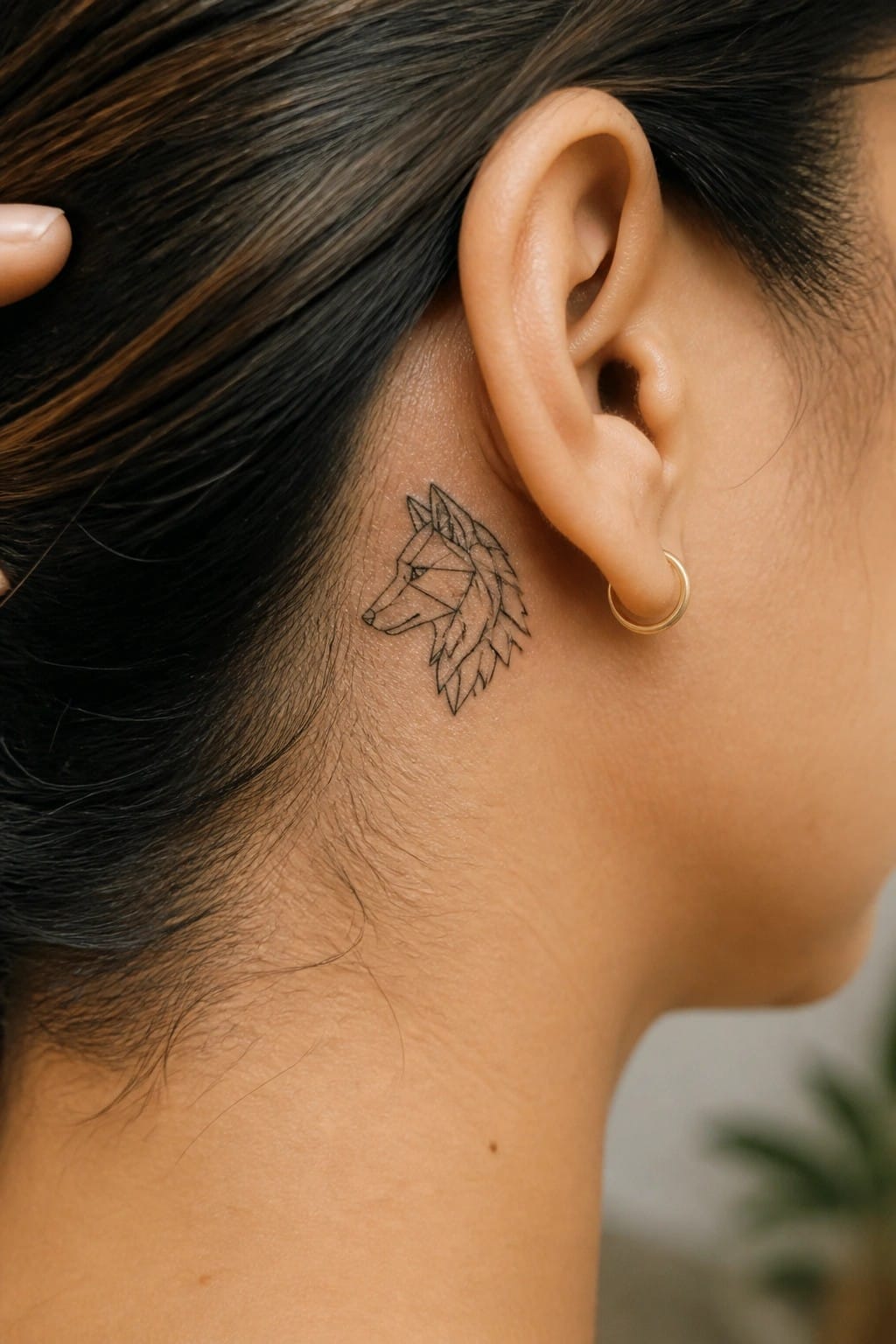 Small geometric wolf tattoo in black line art placed directly behind a woman's ear on the side of the neck.