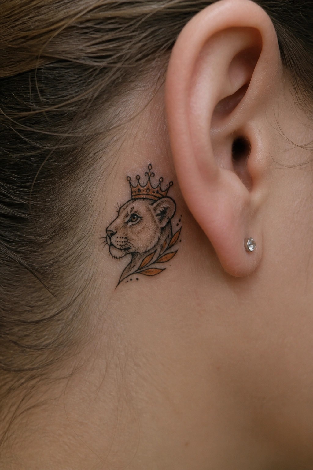 Small neo traditional lioness tattoo with crown and leaves placed behind the ear in profile view