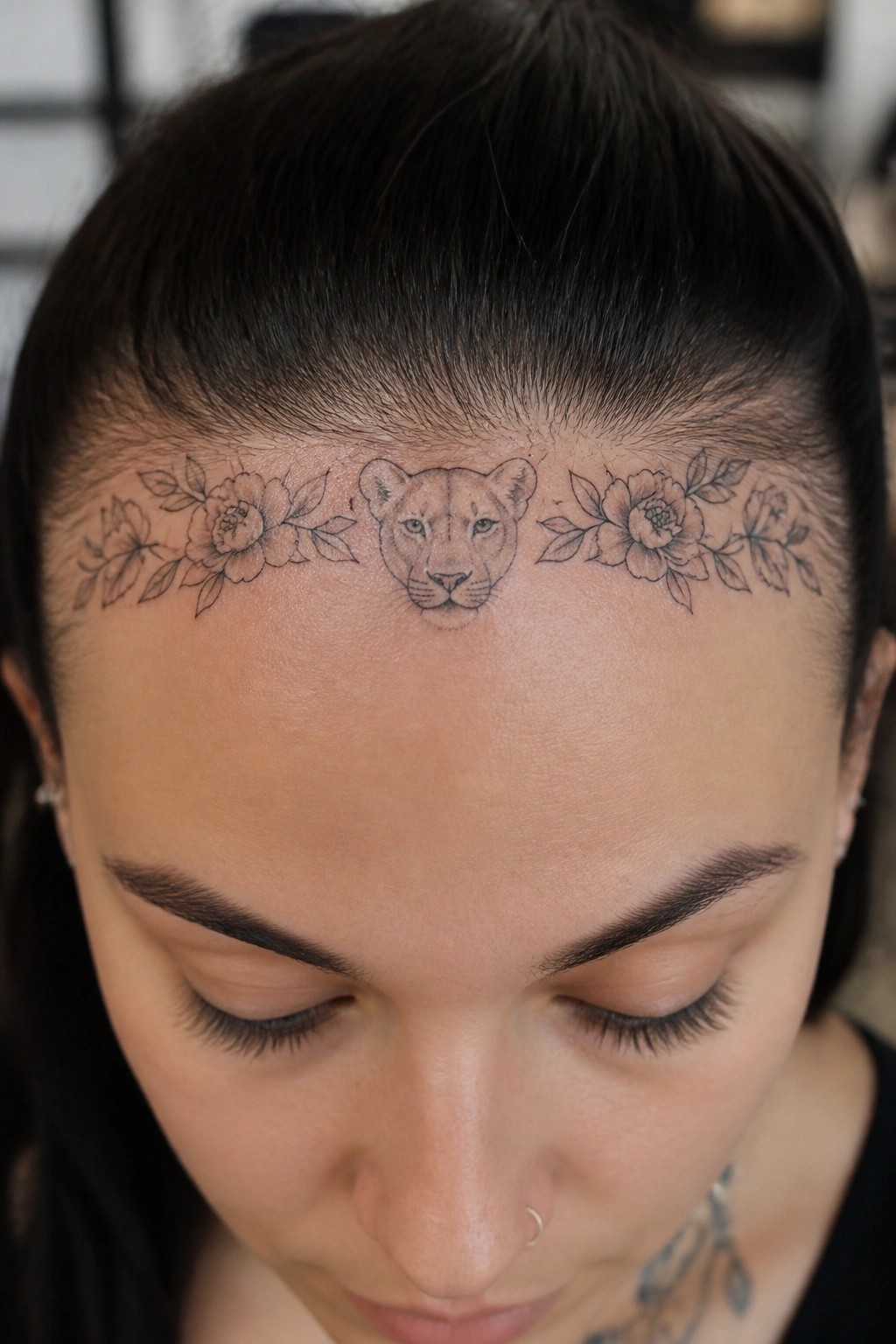 Black line tattoo of a lioness face centered on the forehead with flowers extending to each side just below the hairline