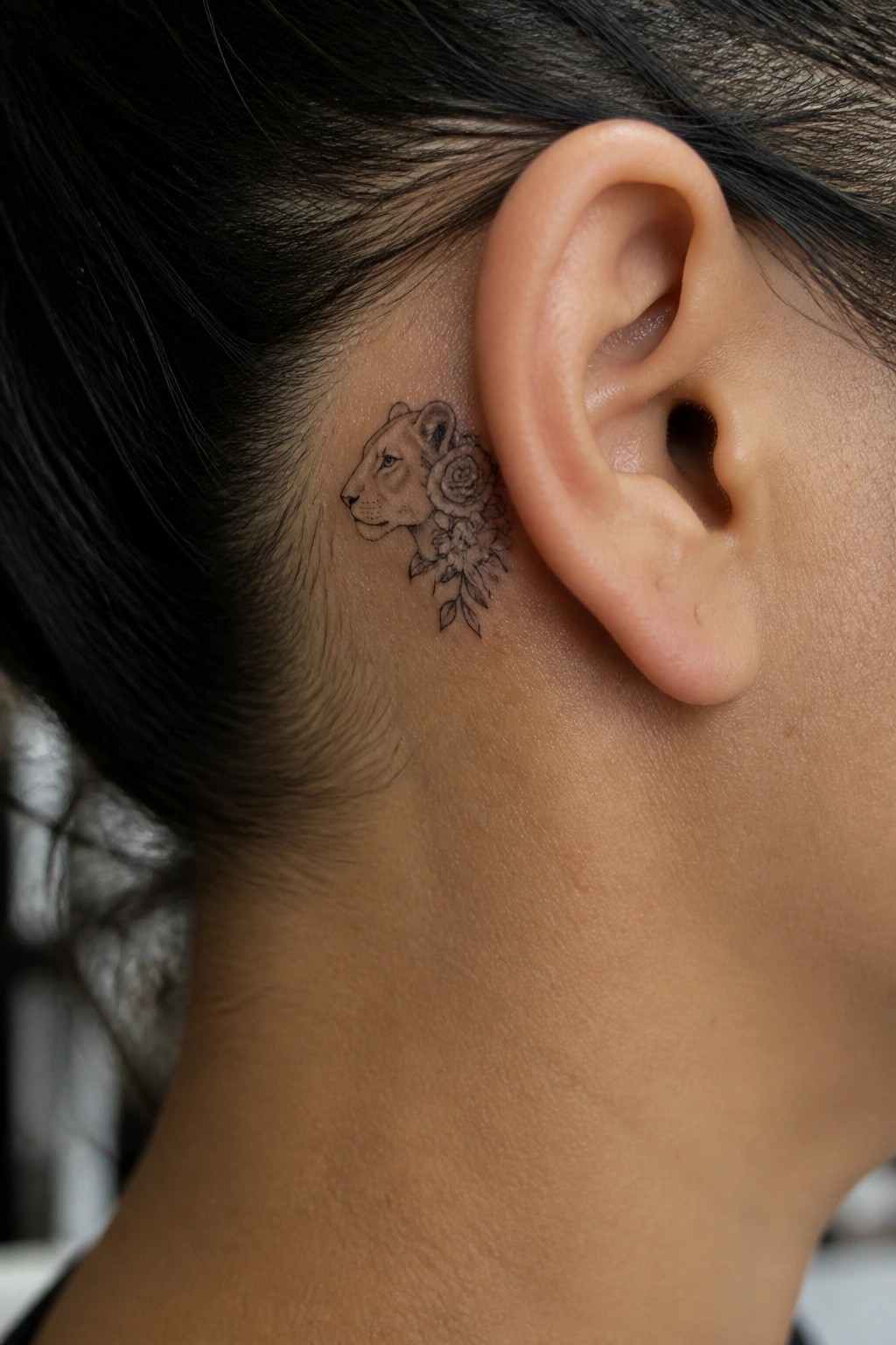 Fine line black and grey tattoo of a lioness in profile with roses and leaves placed directly behind the ear on the side of the head.