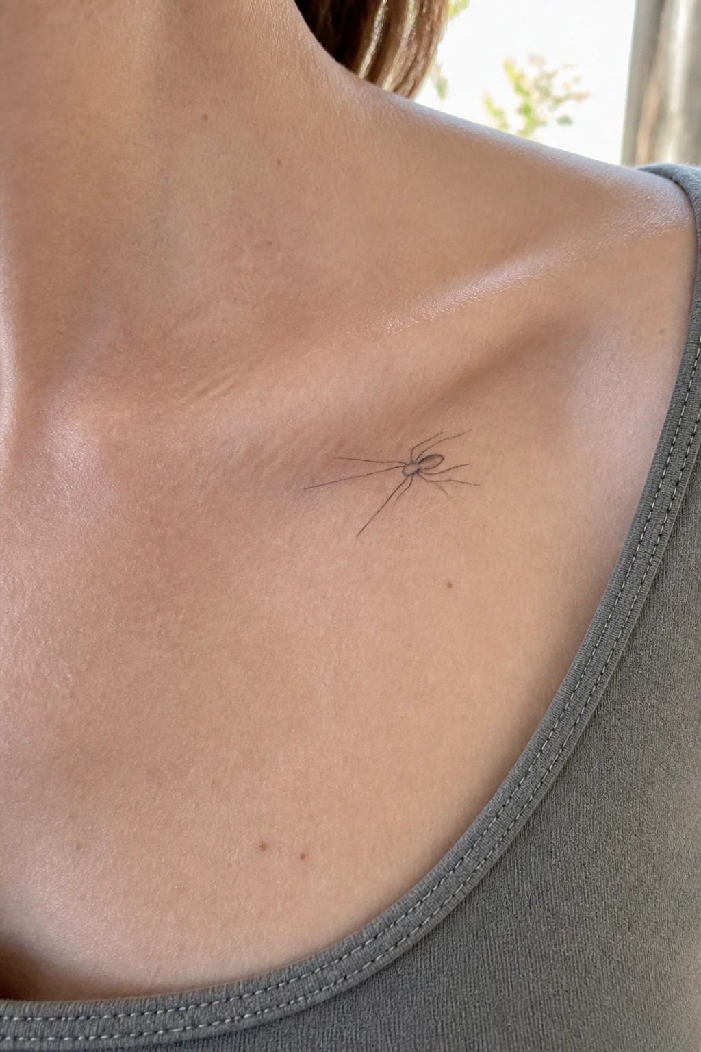 Fine line spider tattoo placed on the collarbone area of a person in a gray top.