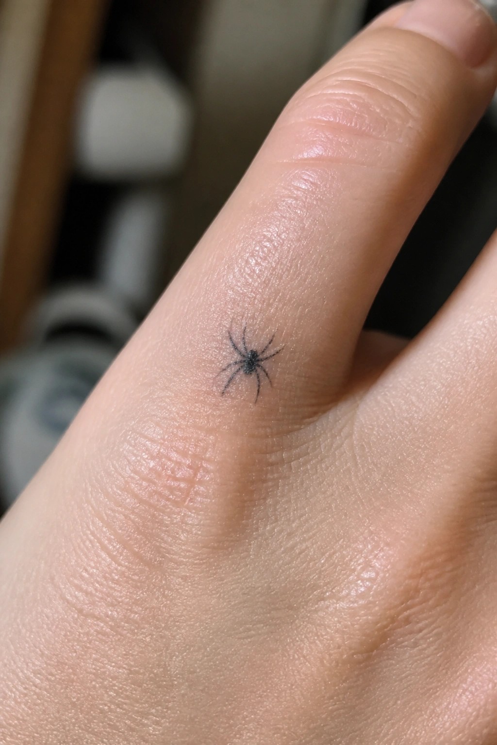 Small black spider tattoo on side of light-skinned finger with creases