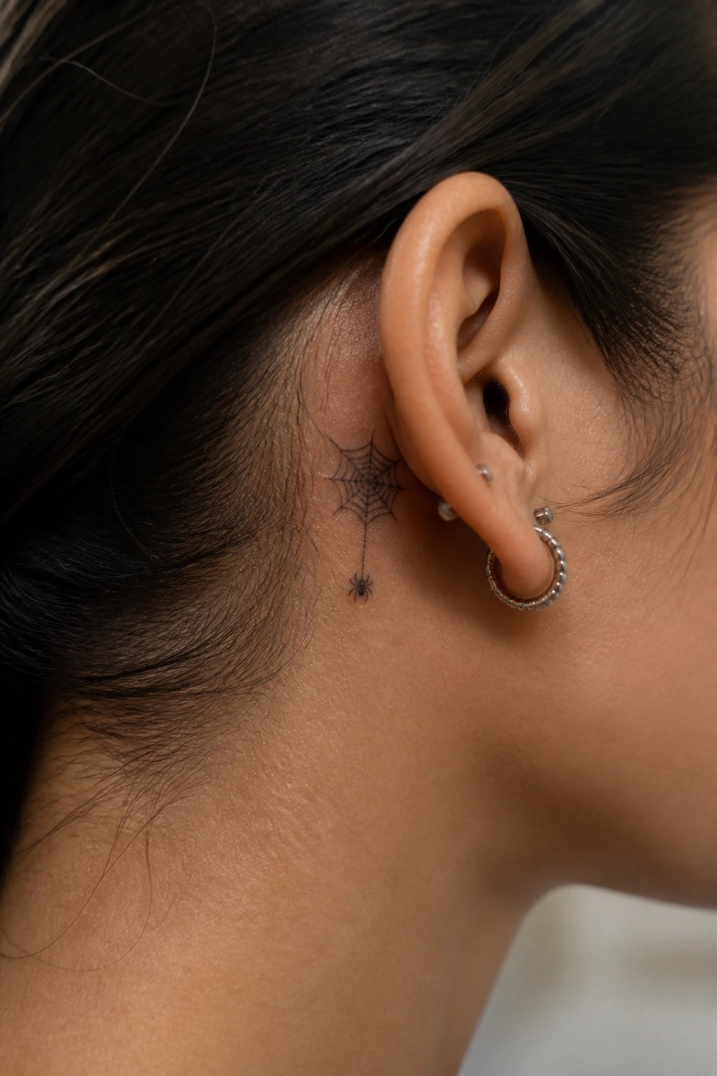 Close-up of ear with black spiderweb tattoo, dangling spider, and silver hoop earring.