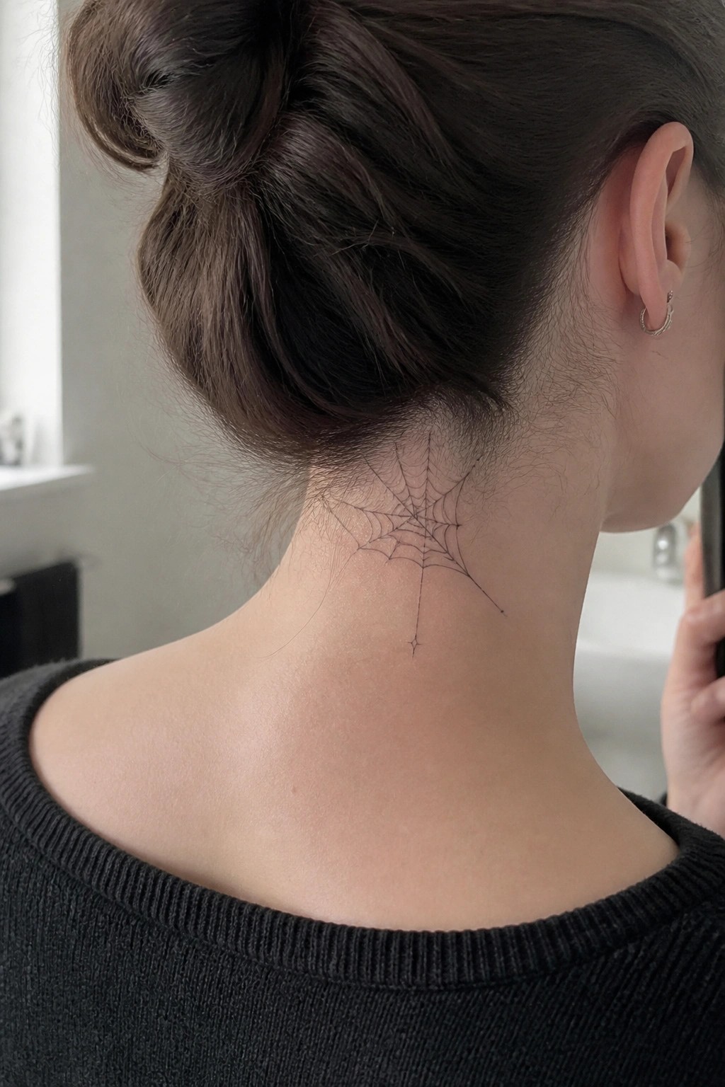 Spiderweb tattoo on neck below dark hair in bun and black sweater