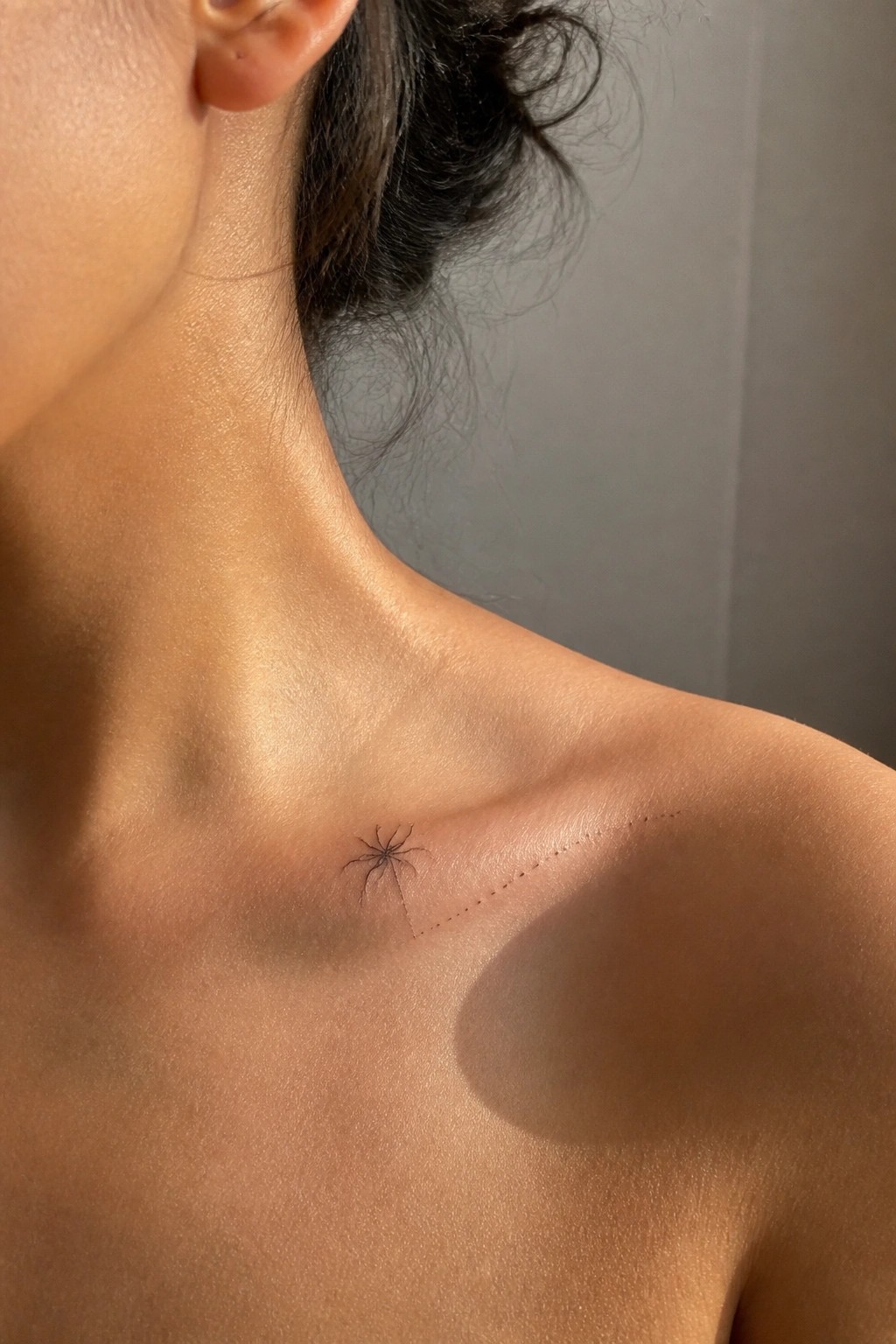 Small black spider tattoo on collarbone with dotted line extending across shoulder.