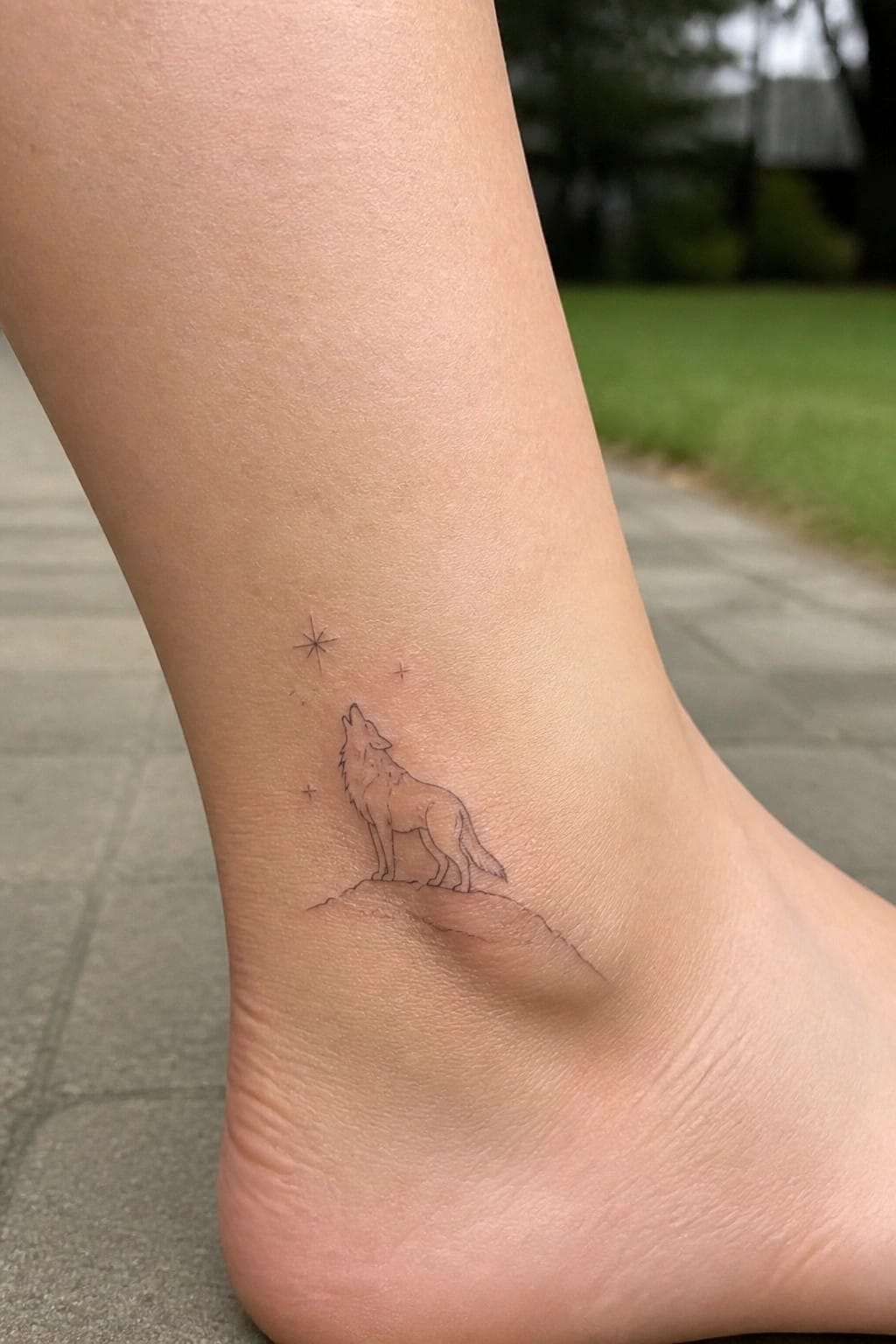 Small line art tattoo of a howling wolf standing on a hill with stars overhead, placed on the ankle.