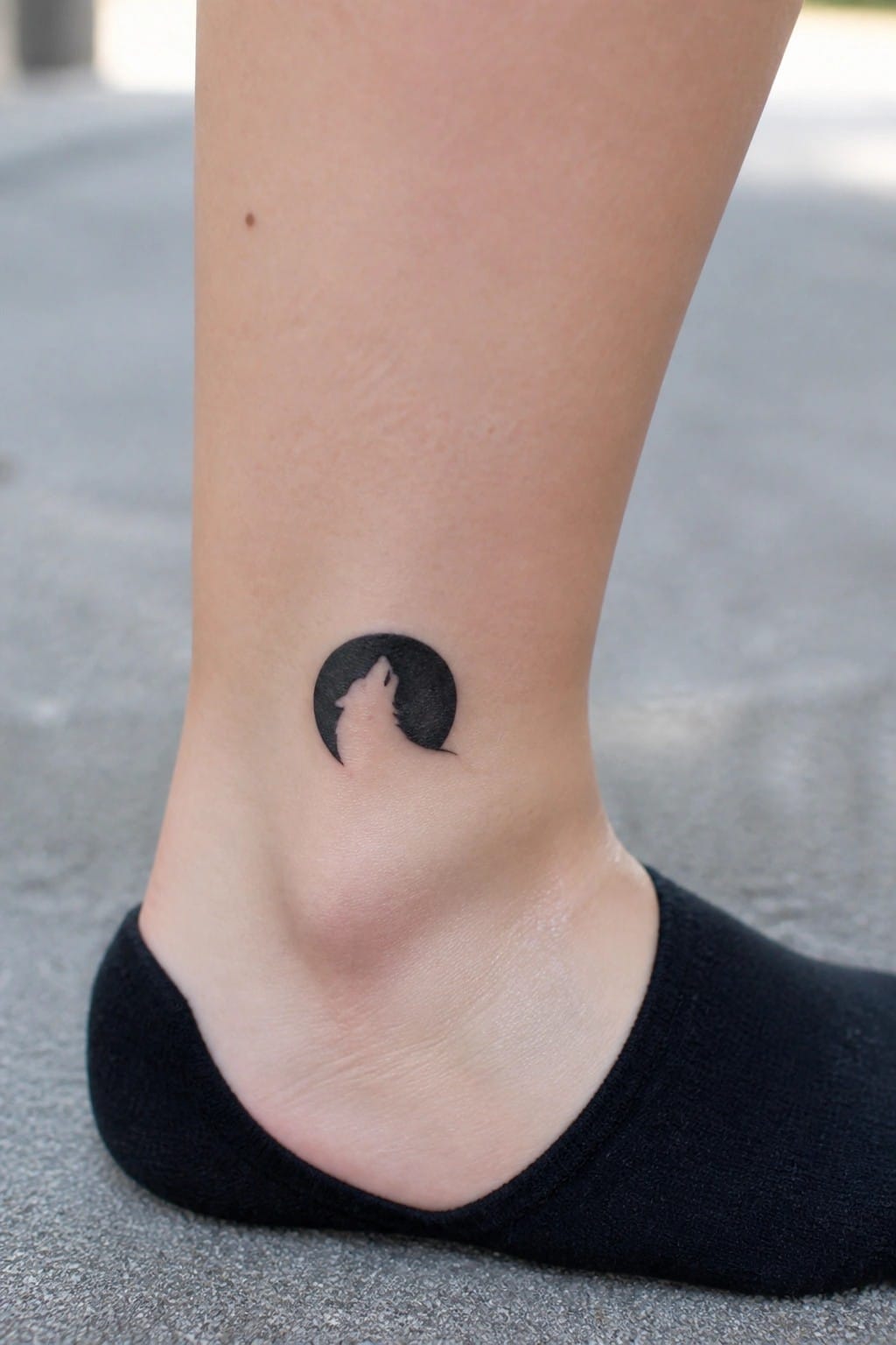 Small black circular tattoo of a wolf howling on the ankle above a black sock.