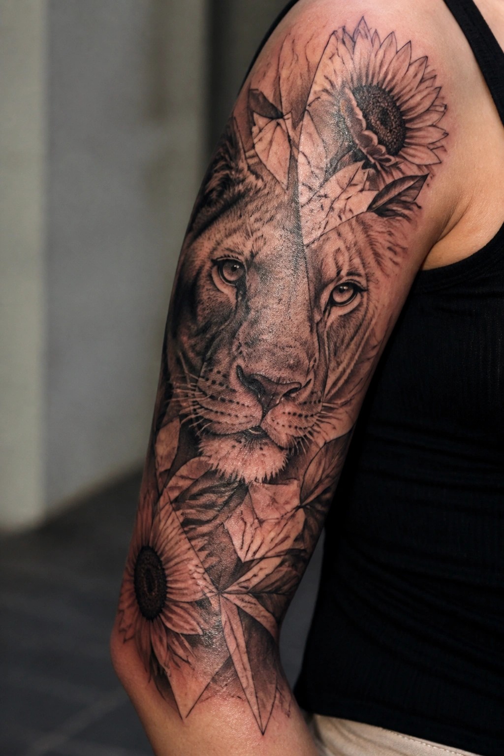 Realistic black and grey lioness face tattoo on upper arm with sunflowers and geometric leaf patterns integrated around it