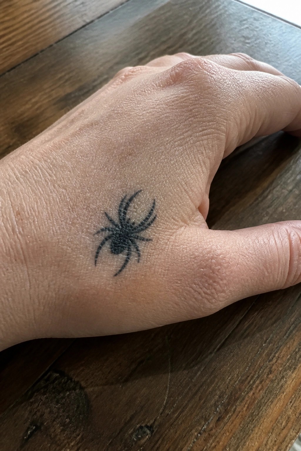 Small blackwork spider tattoo on the back of a hand near the thumb base