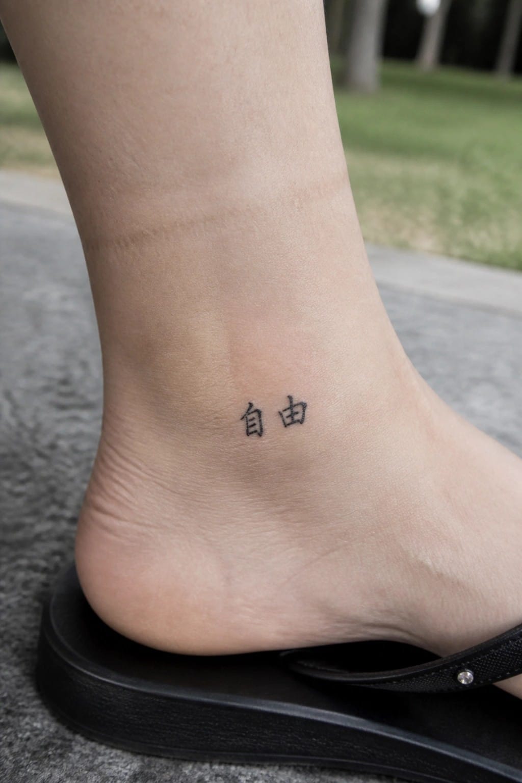Small Chinese characters spelling freedom tattooed on the outer ankle above a foot wearing a black flip-flop
