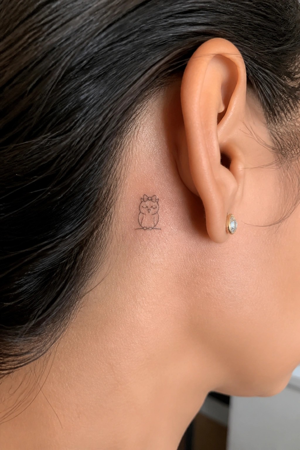 Small owl tattoo with bow behind ear amid dark hair and gold stud earring.