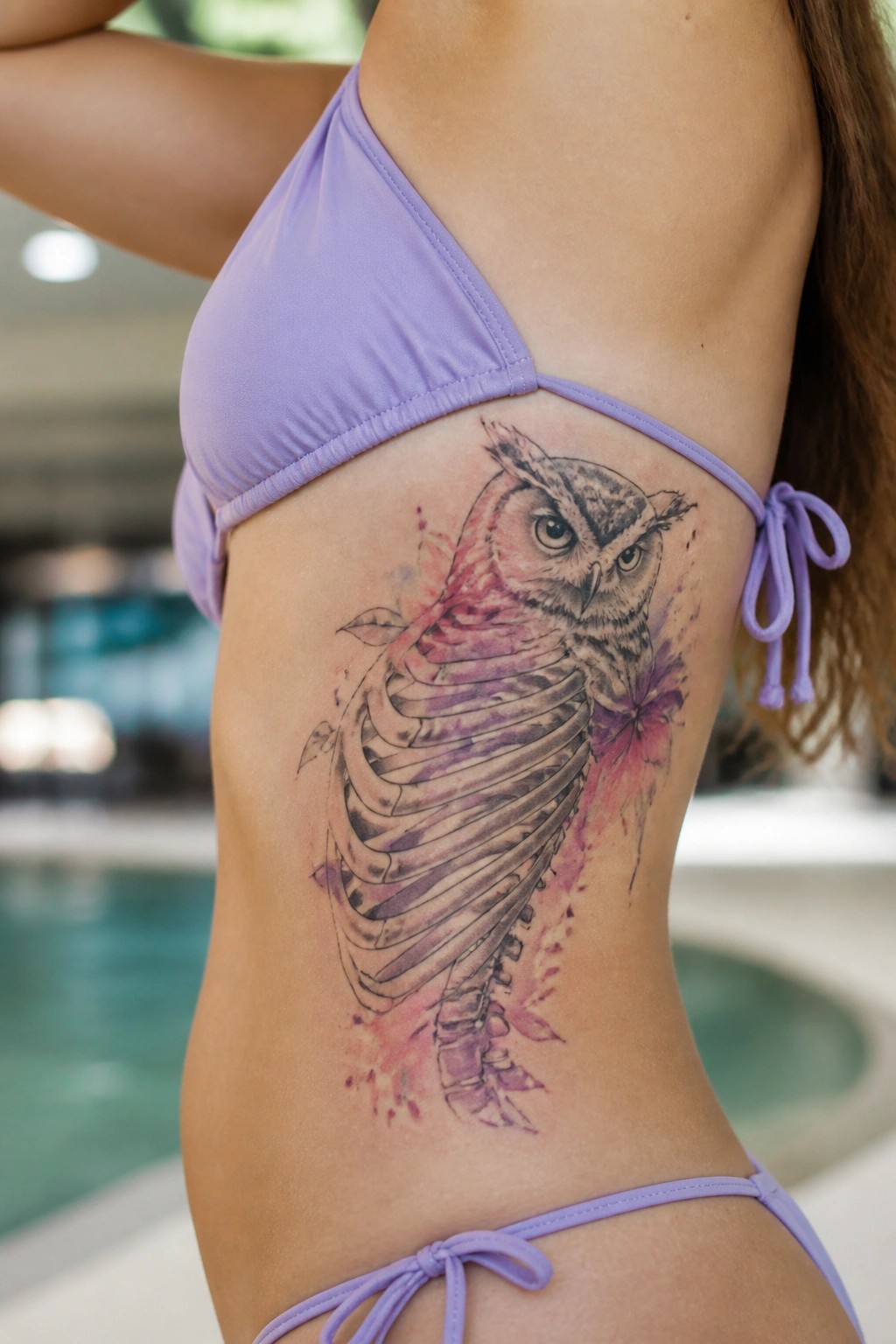 A large owl tattoo on a woman's side torso blending ribcage bones with watercolor pink and purple accents, shown on skin next to a purple bikini top.