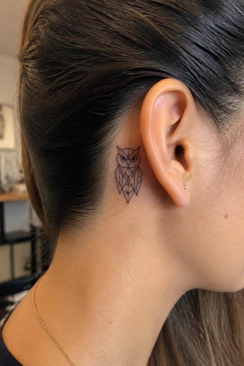 Small geometric owl tattoo in black line art placed behind a woman's ear on the side of her head.