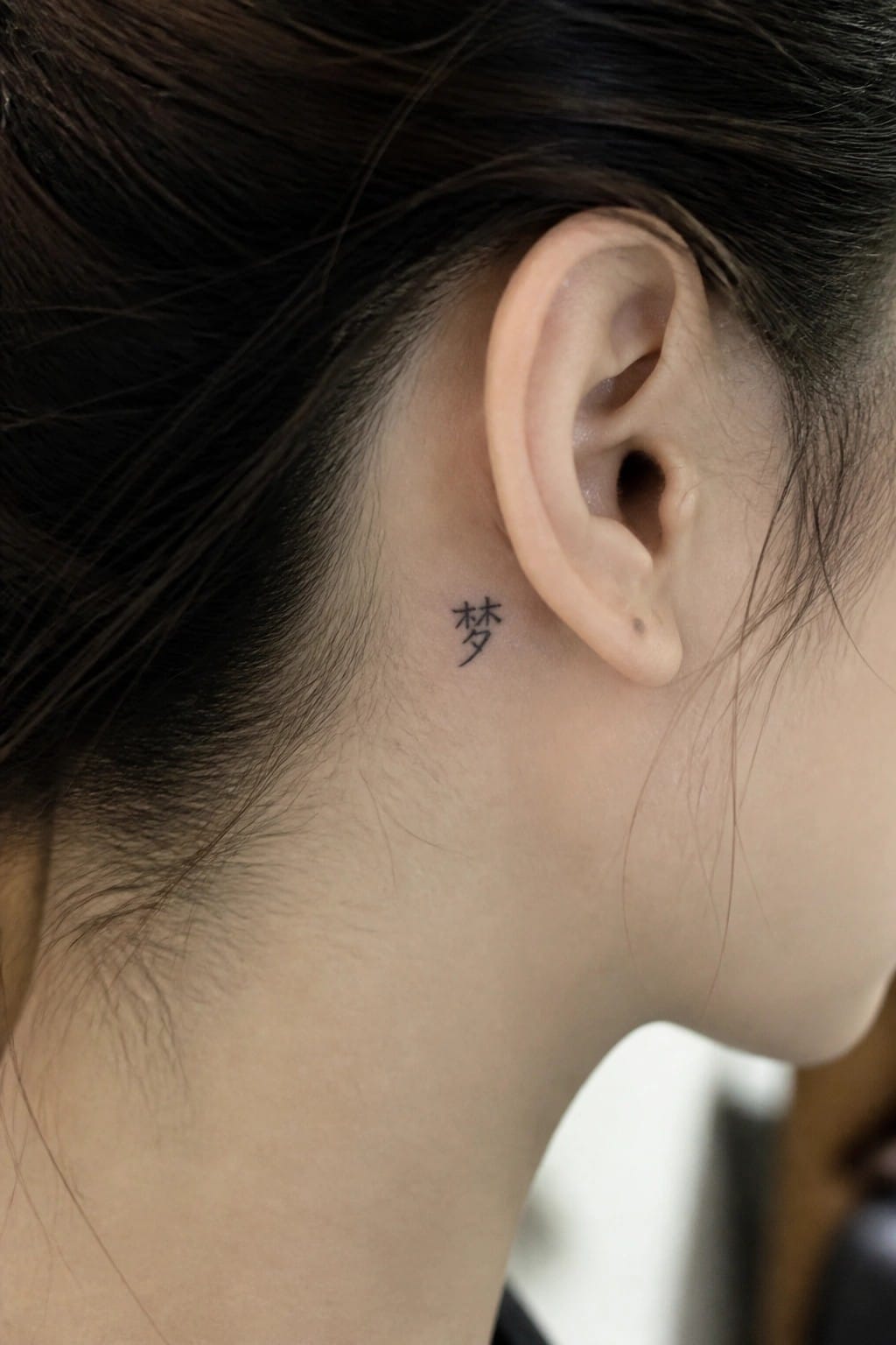 Small black Chinese character tattoo behind person's ear amid dark hair
