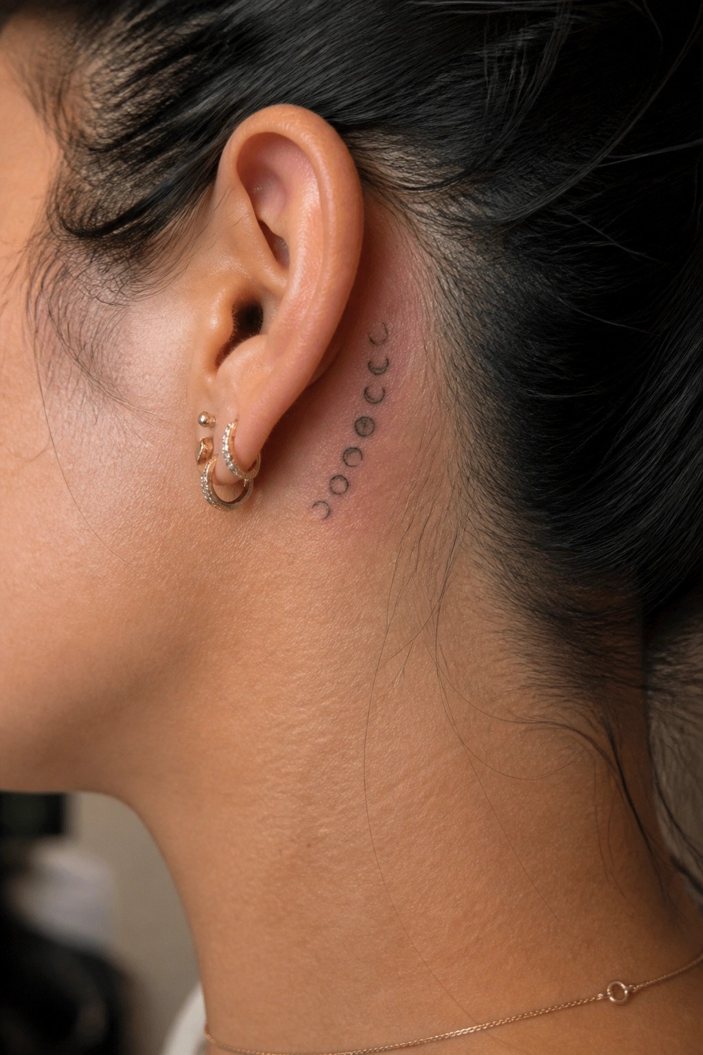 Ear with moon phase tattoo, gold hoop earrings, dark pulled-back hair, thin necklace
