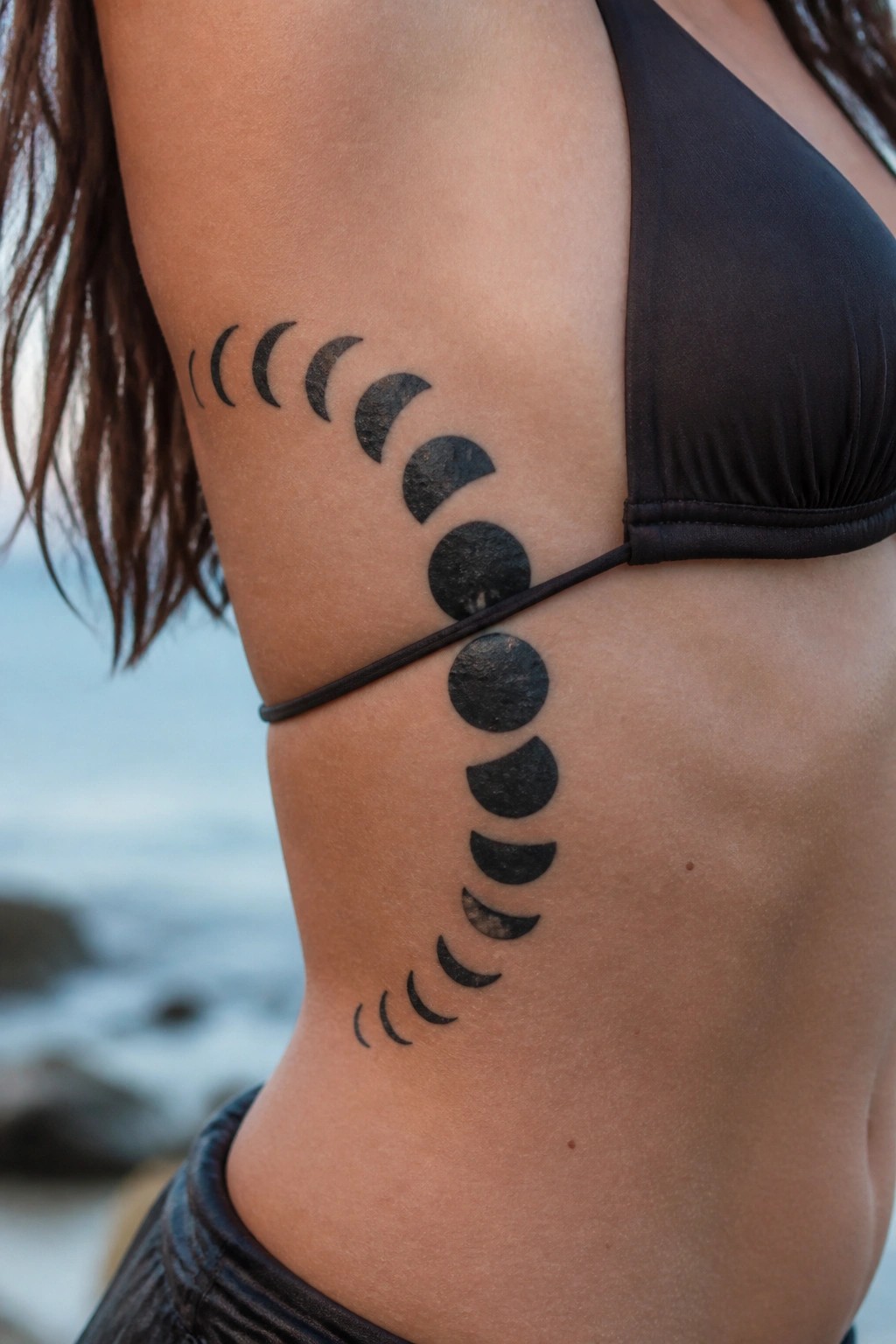 Black ink moon phase tattoo curving along a woman's side torso from ribs to waist showing the full lunar cycle.