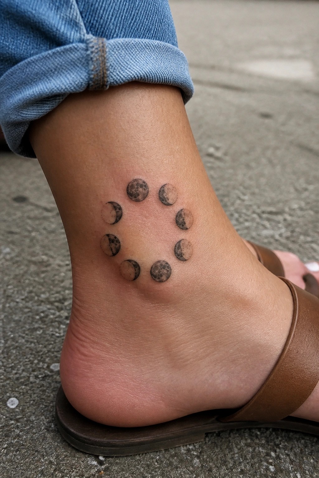 Small circular moon phase tattoo on the ankle in black and gray ink showing the full lunar cycle.