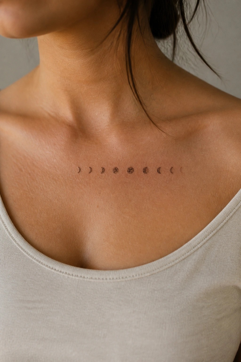 Moon phase tattoo across collarbone above light beige tank top.