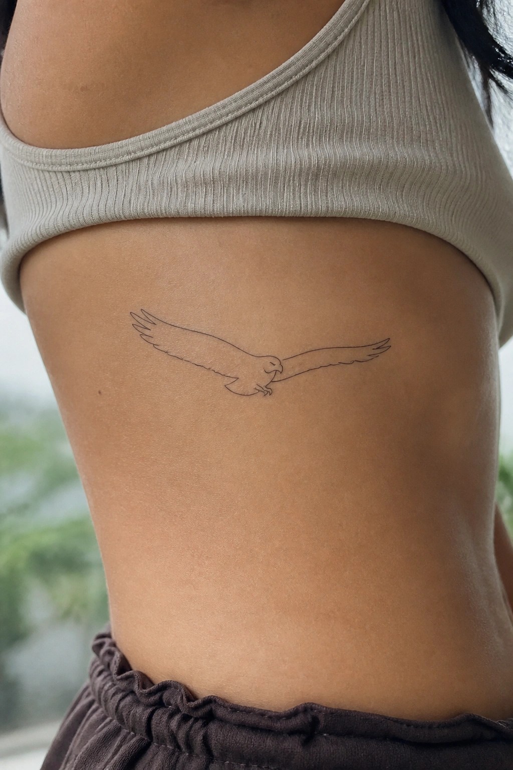Minimalist black line tattoo of flying eagle on ribcage below gray crop top
