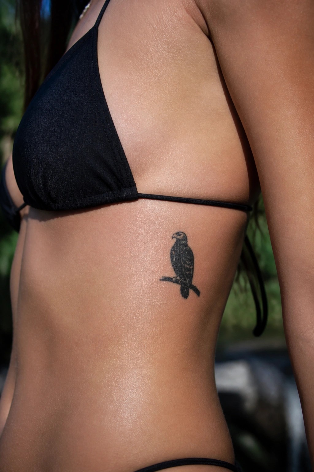 Small black and grey eagle tattoo perched on a branch, placed on the side torso under a bikini strap.