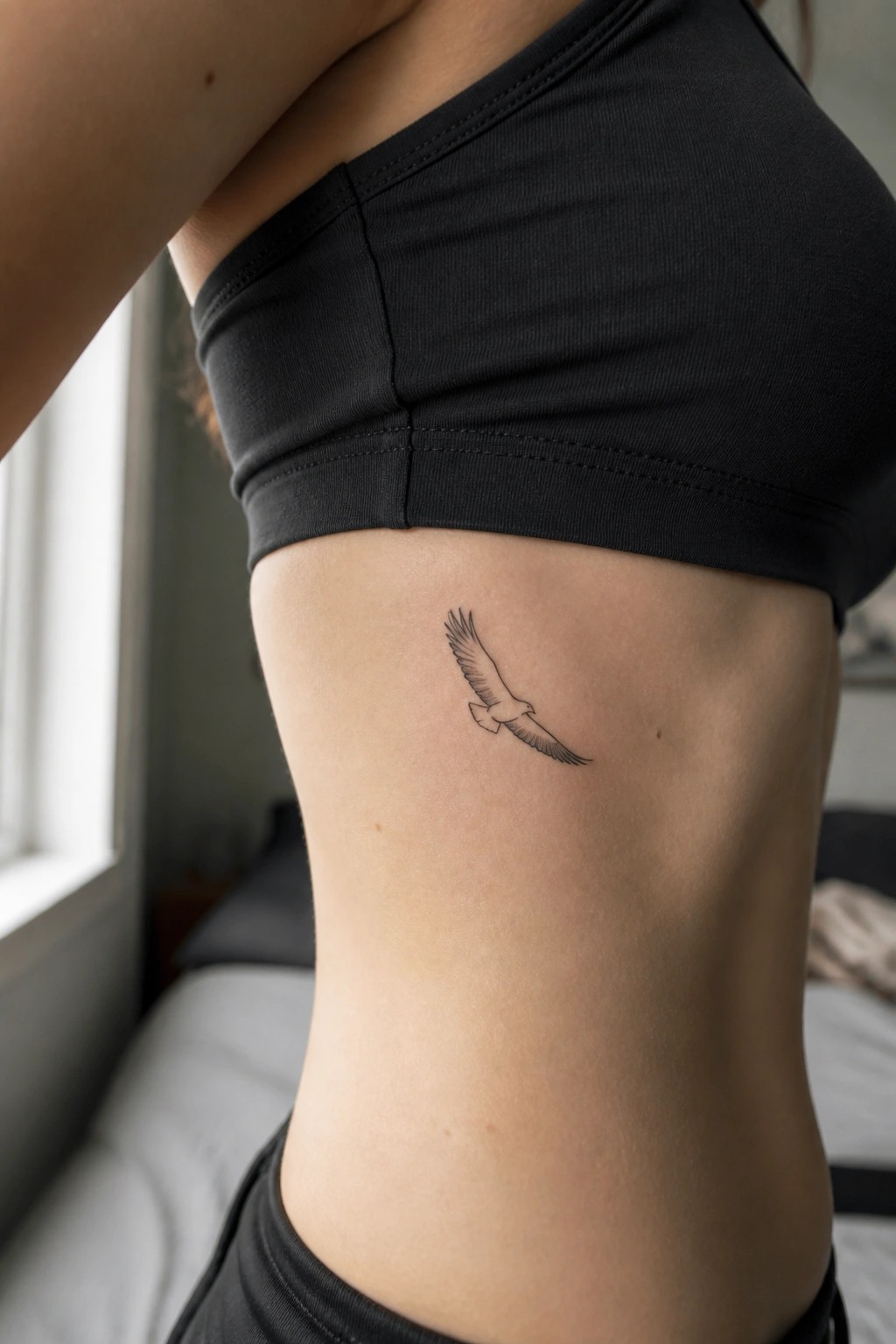 Small black line art eagle tattoo in flight on the side torso below a black crop top.