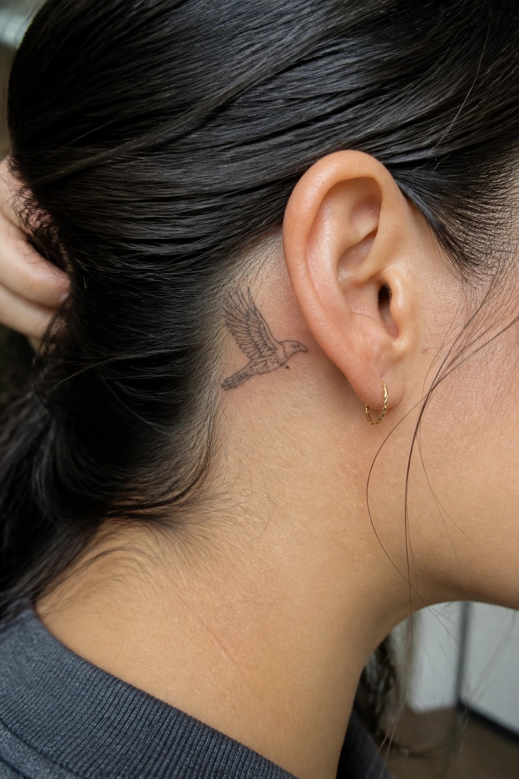 Dark-haired person with bird tattoo behind ear and small gold hoop earring