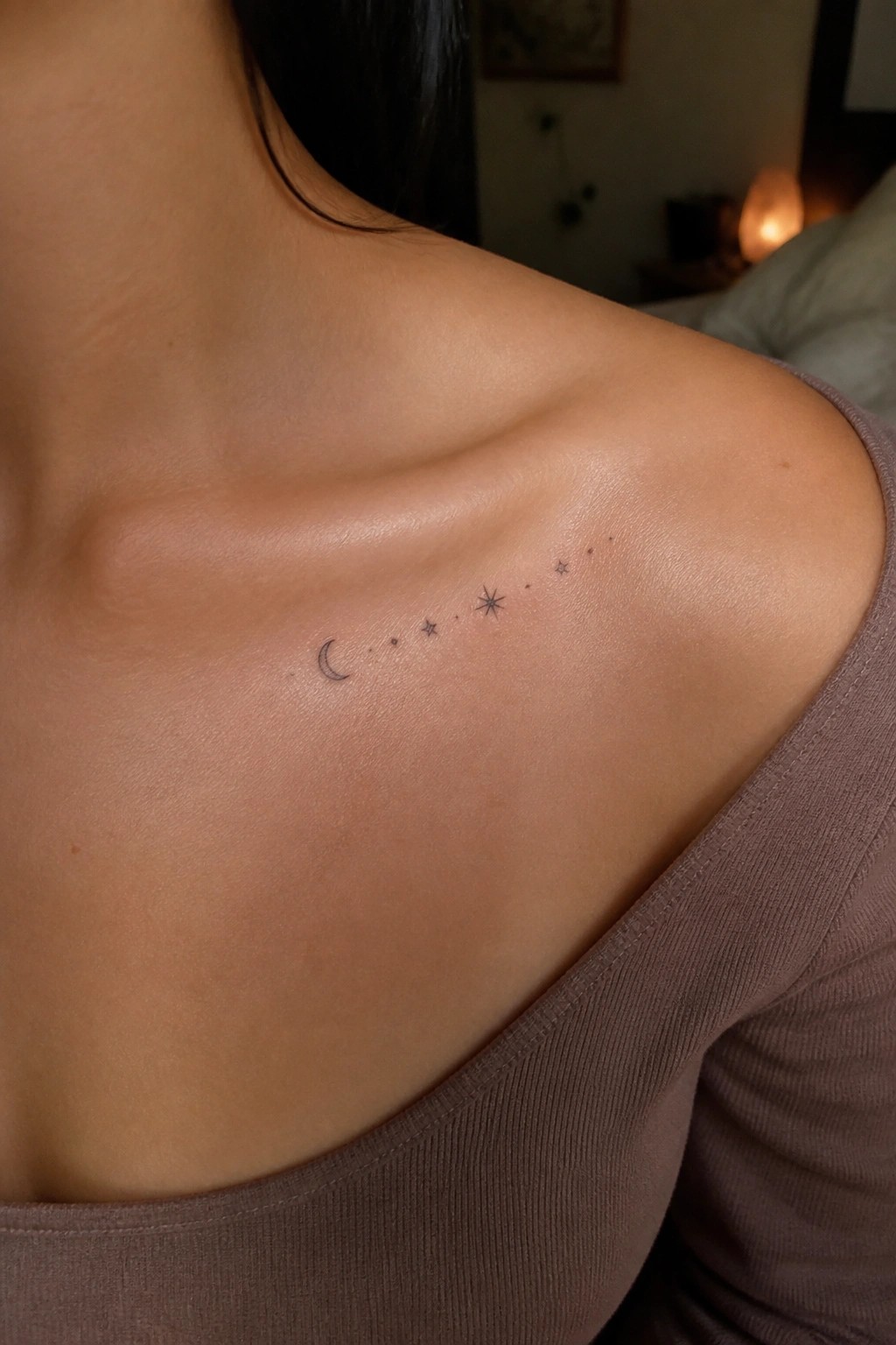 Fine line tattoo of a crescent moon followed by dots and stars across a woman's shoulder near the collarbone.