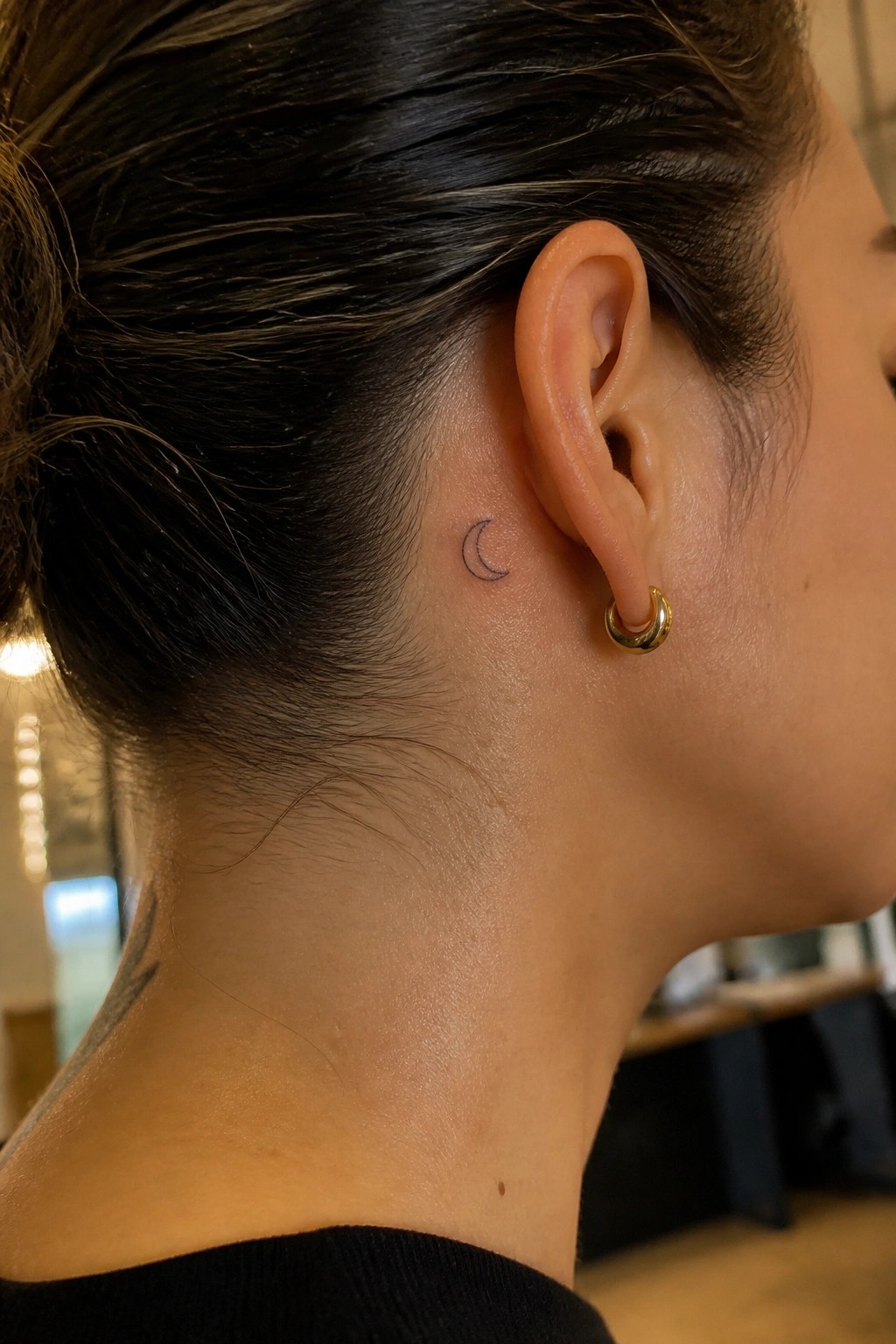 Dark-haired woman with crescent moon tattoo behind ear and gold hoop earring.