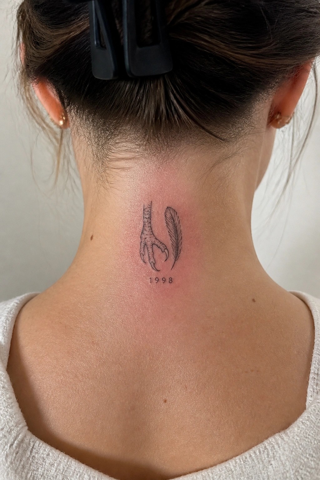 Small black line tattoo of an eagle claw and feather on the nape of the neck with 1998 below it