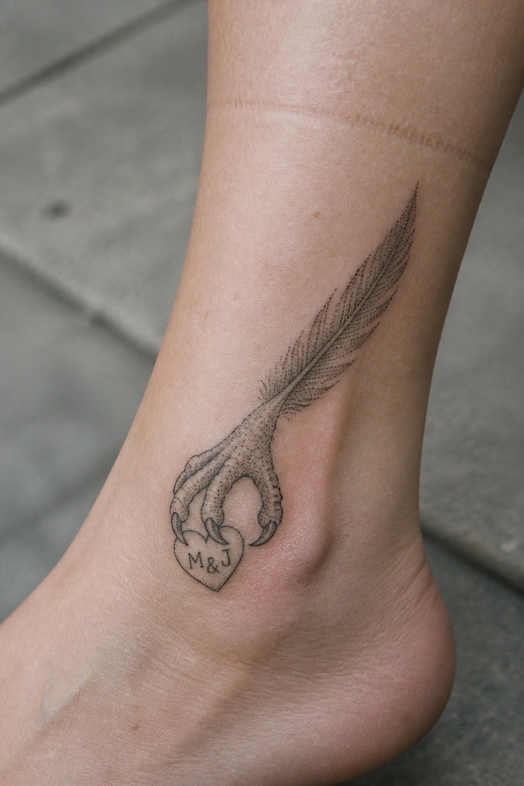 Black ink eagle claw tattoo holding a heart with M and J initials on a woman's ankle, with a long feather extending upward.