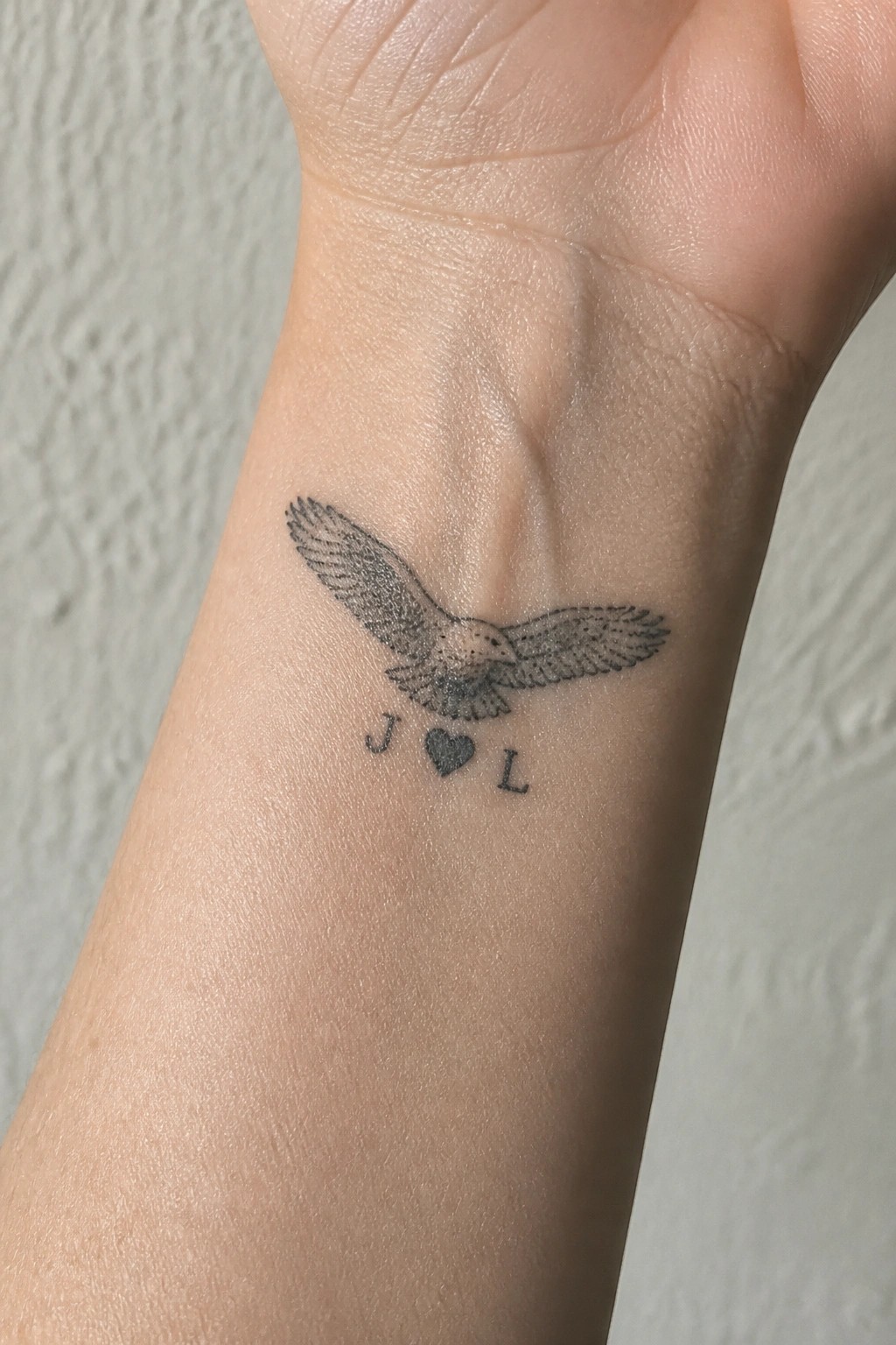 Black ink flying bird tattoo on wrist above J heart L
