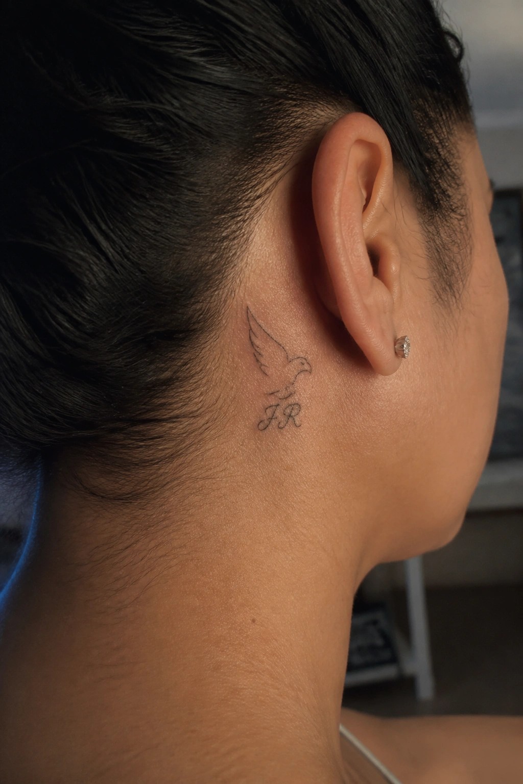 Dark hair in bun reveals bird tattoo with JR behind ear and diamond stud.