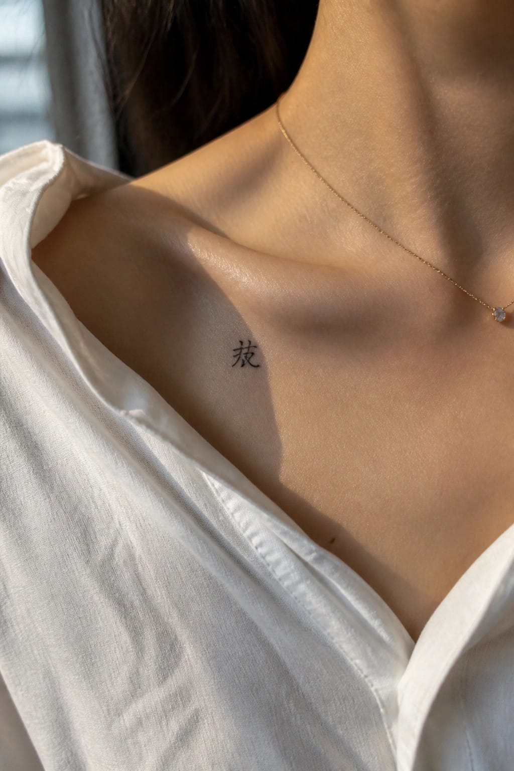 Collarbone with small black Chinese tattoo, thin gold necklace, and white shirt in sunlight