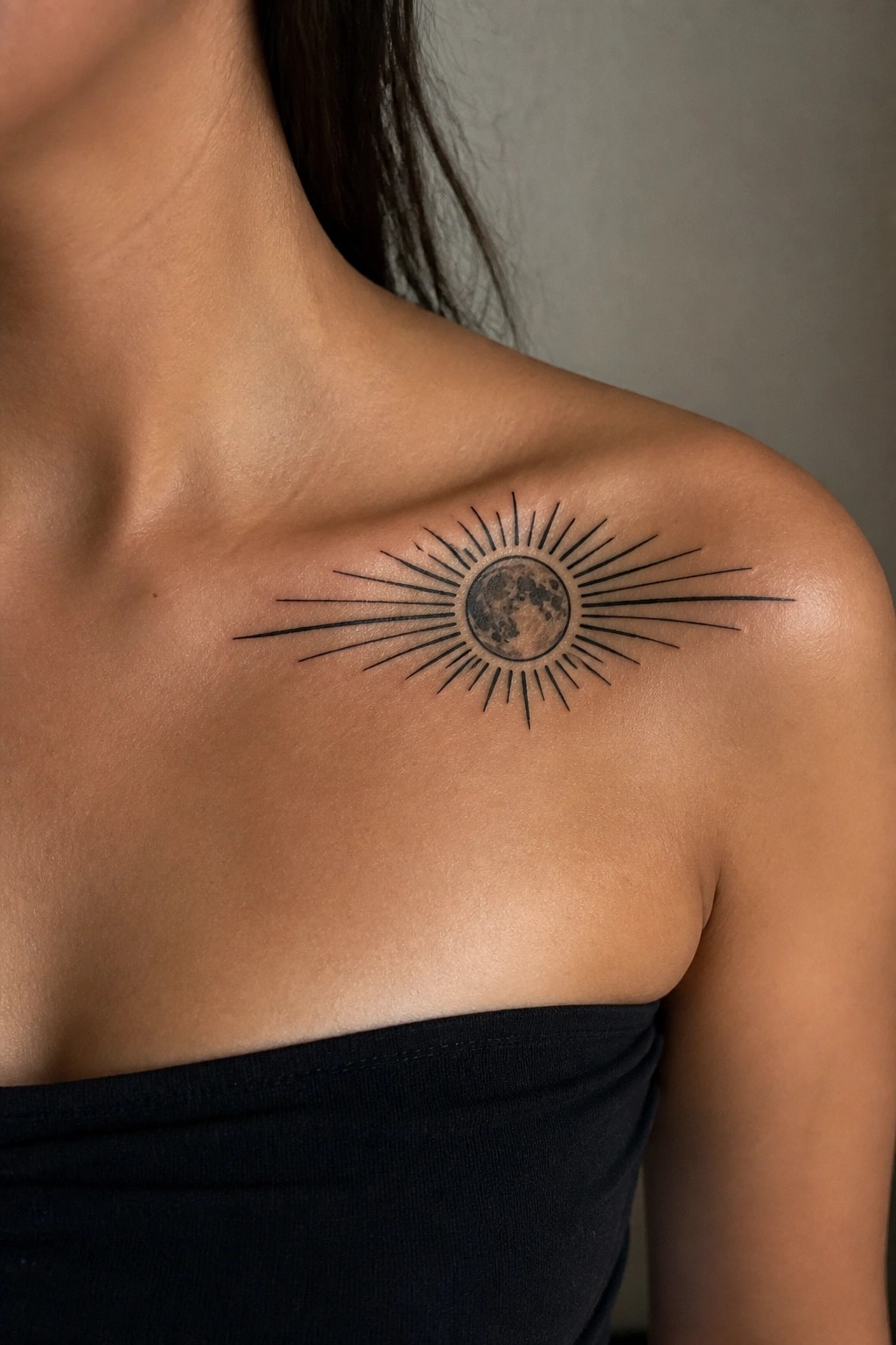 Black ink tattoo of a full moon with radiating lines placed on a woman's shoulder near the collarbone