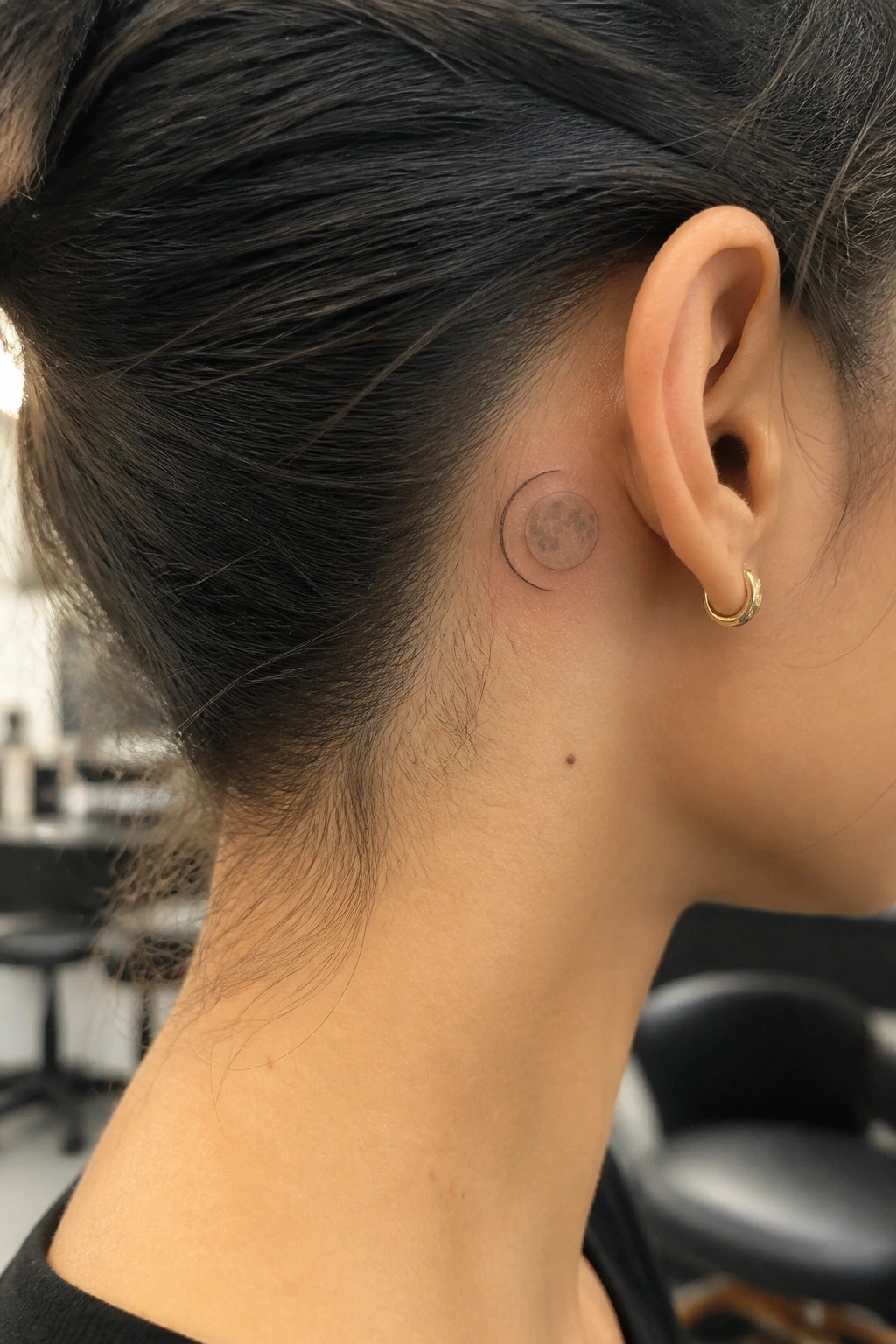 Fine line tattoo of a crescent moon overlapping a shaded full moon placed behind the ear on the side of the head